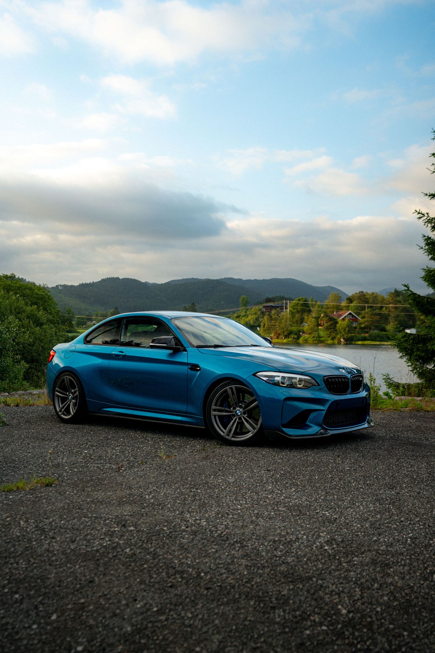 BMW M2 in bright blue metallic paint parked against scenic mountain lake backdrop.