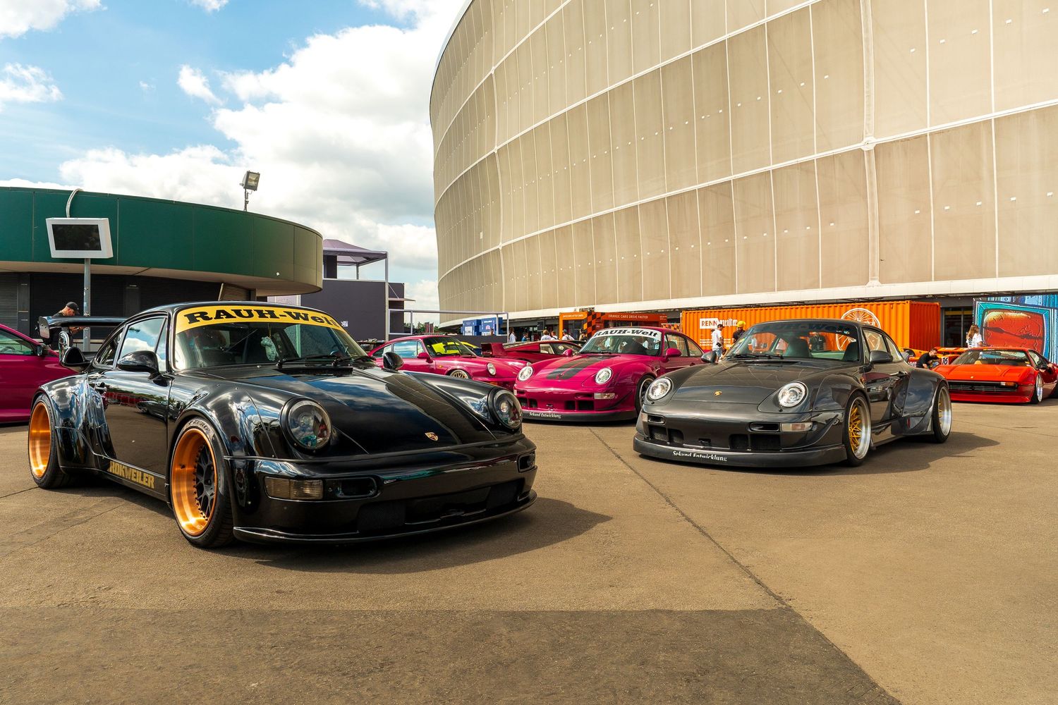 Two modified Porsche sports cars parked at a car show venue, one black and one silver, with custom wheels and racing styling.