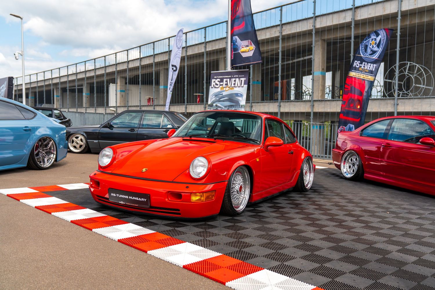 Classic red Porsche 911 displayed at a car show on a checkered floor mat with other vehicles in background.