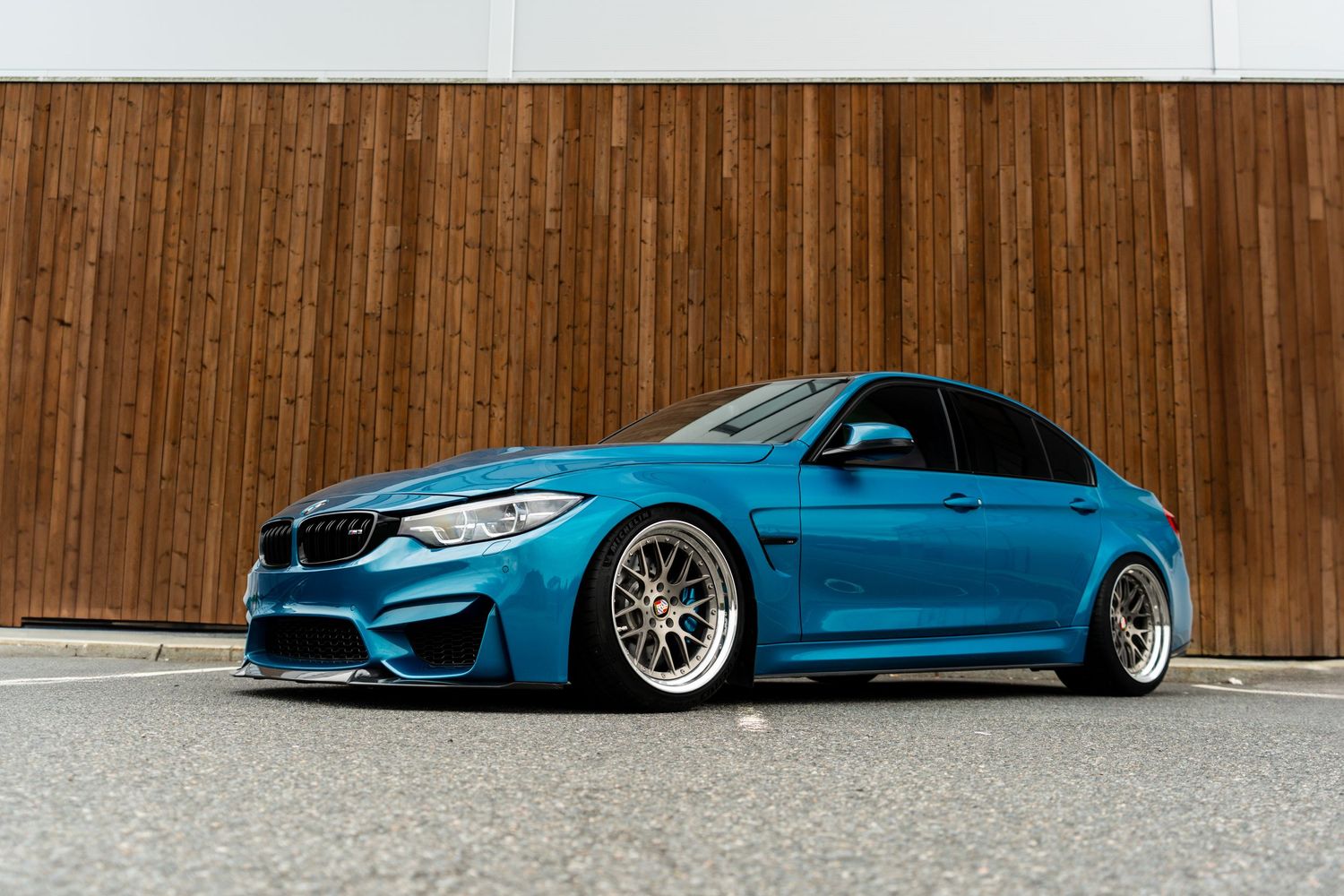Modified BMW M3 in metallic blue paint sitting low with aftermarket wheels against wooden wall.