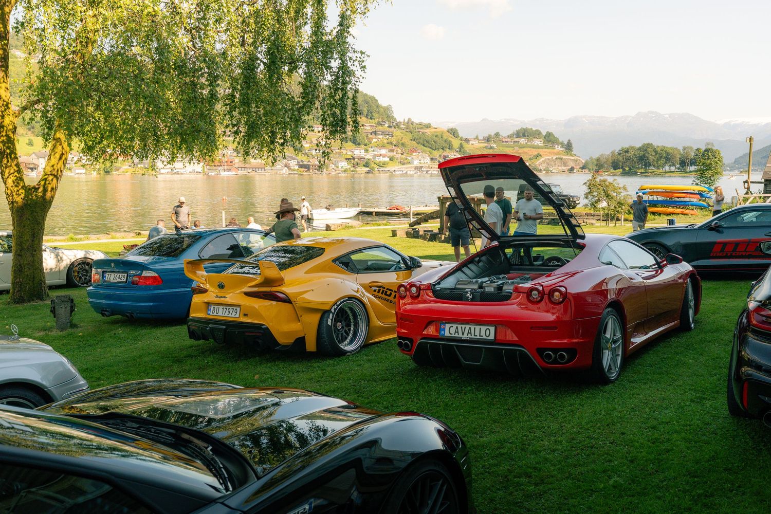 Red Ferrari with open rear hood, yellow Toyota Supra and blue BMW parked on a lakeside lawn at an outdoor car show.