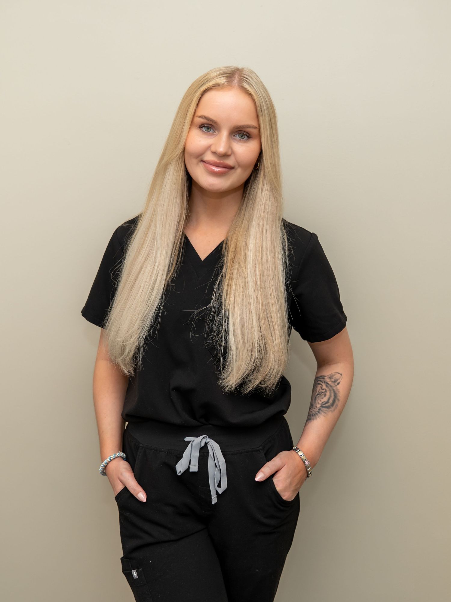 Smiling woman with long blonde hair wearing black scrubs, standing with hands in pockets, with a forearm tattoo, against a neutral wall.