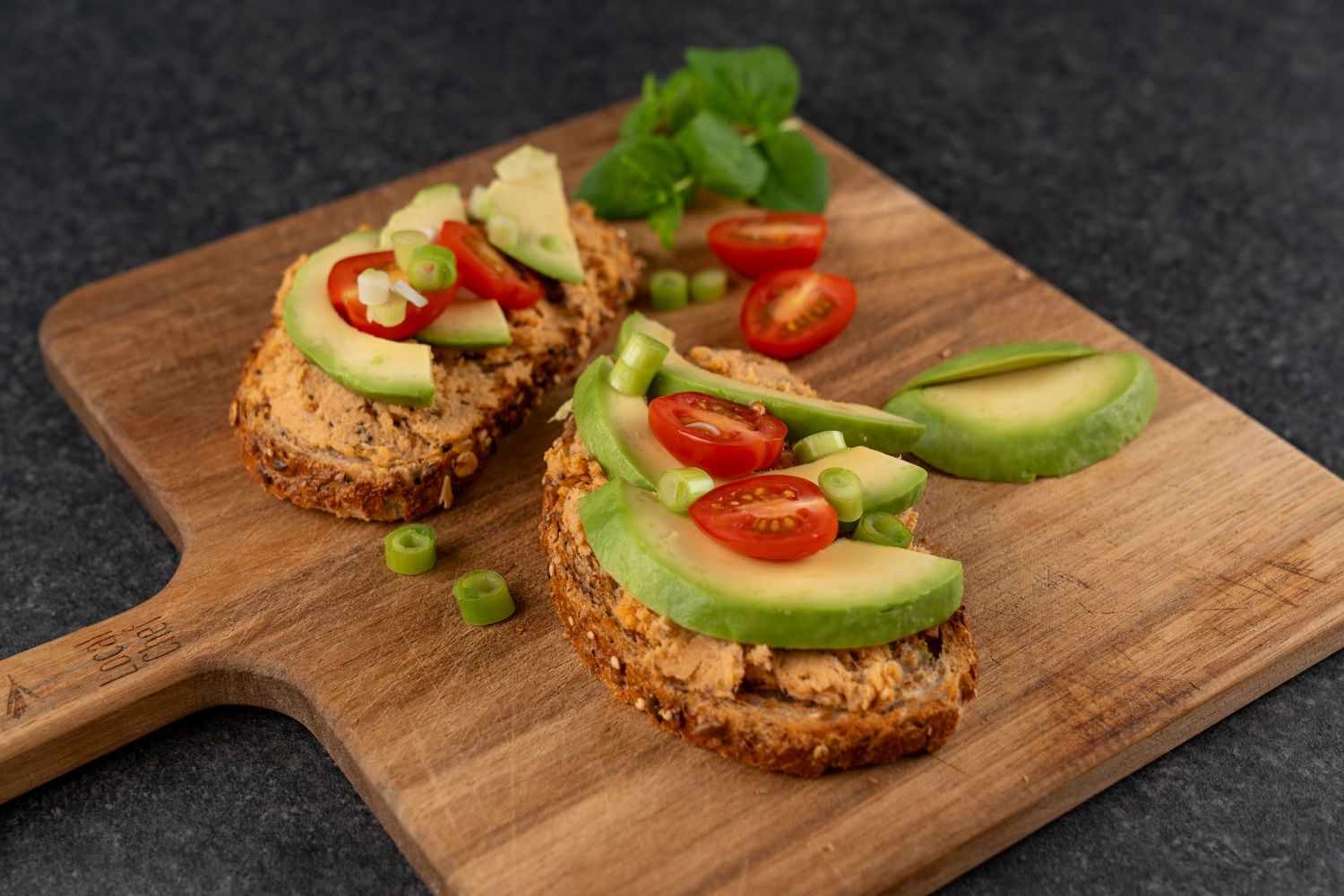 Two open-faced toasts topped with hummus, sliced avocado, cherry tomato slices and green onion, served on a wooden board with fresh herbs.