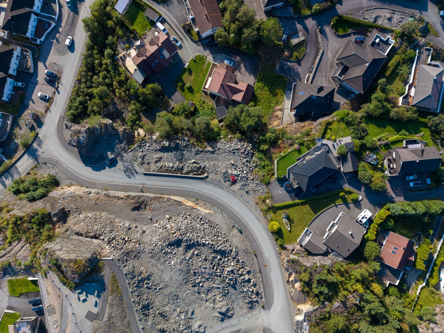 Aerial view of a curving road winding past a rocky excavated lot and modern hillside homes surrounded by trees.