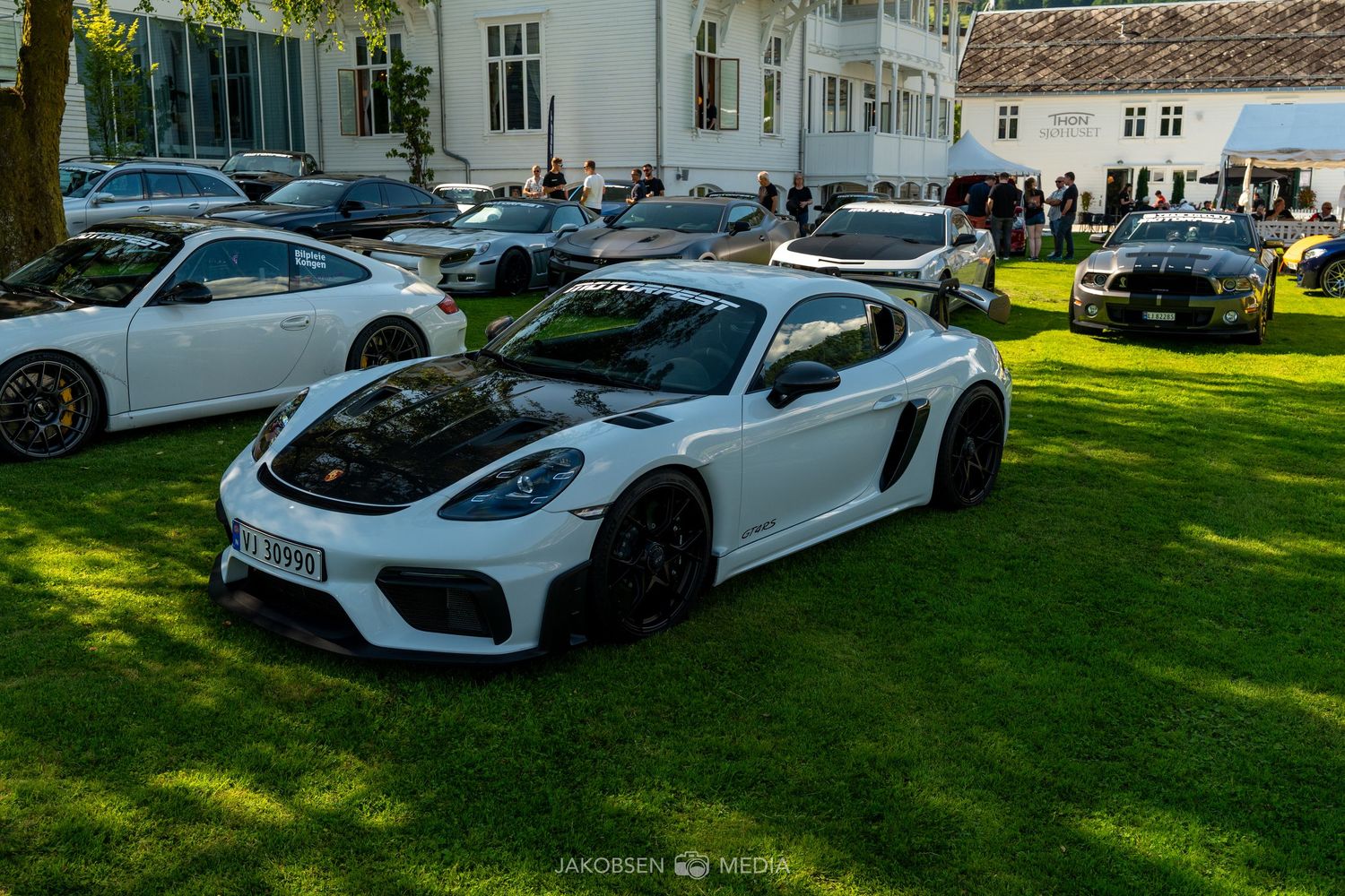 A white Porsche sports car displayed on a manicured lawn with other luxury vehicles in the background.