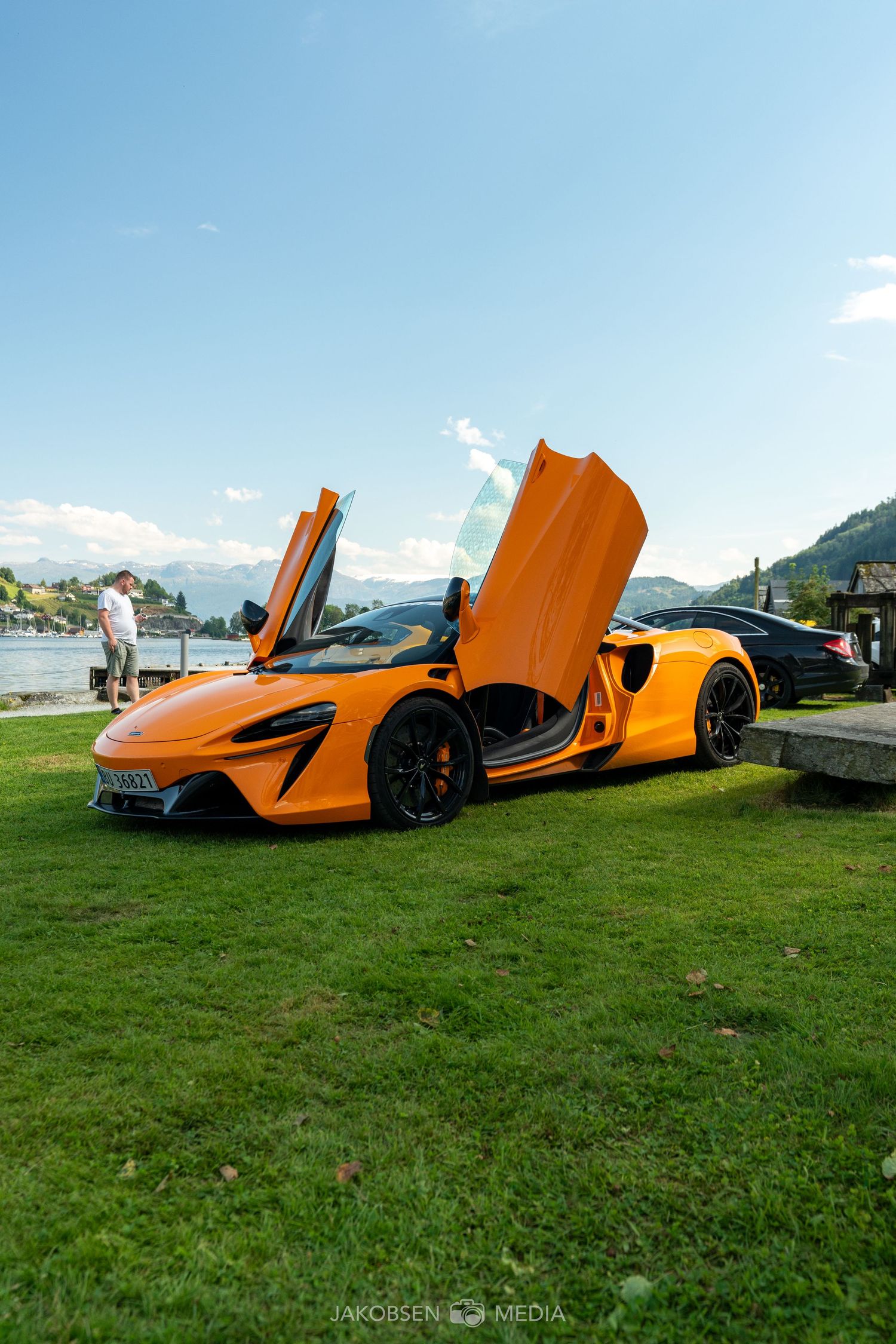 Orange McLaren with dramatic butterfly doors open displayed on grass with other supercars.