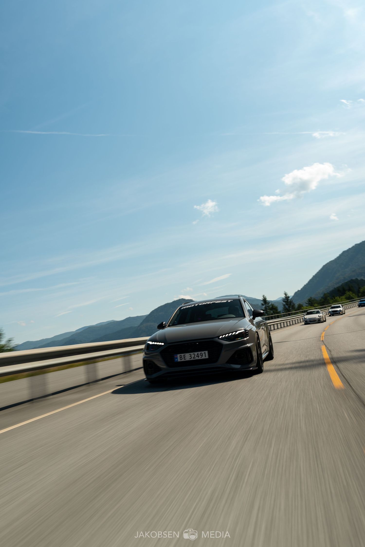 Sports cars driving in formation on a scenic mountain highway under blue skies.