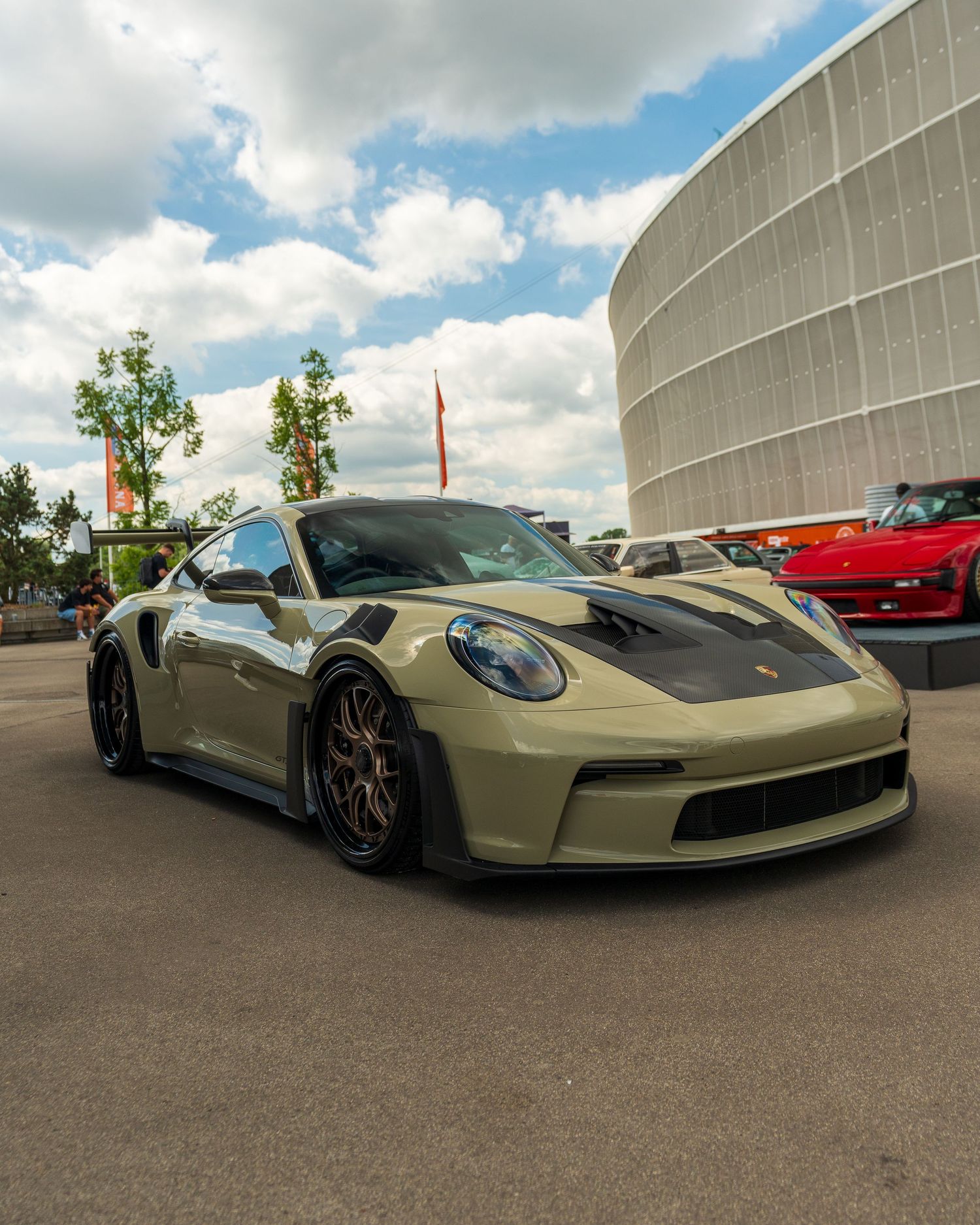 Military green Porsche 911 GT3 parked outside a modern curved building under cloudy skies.
