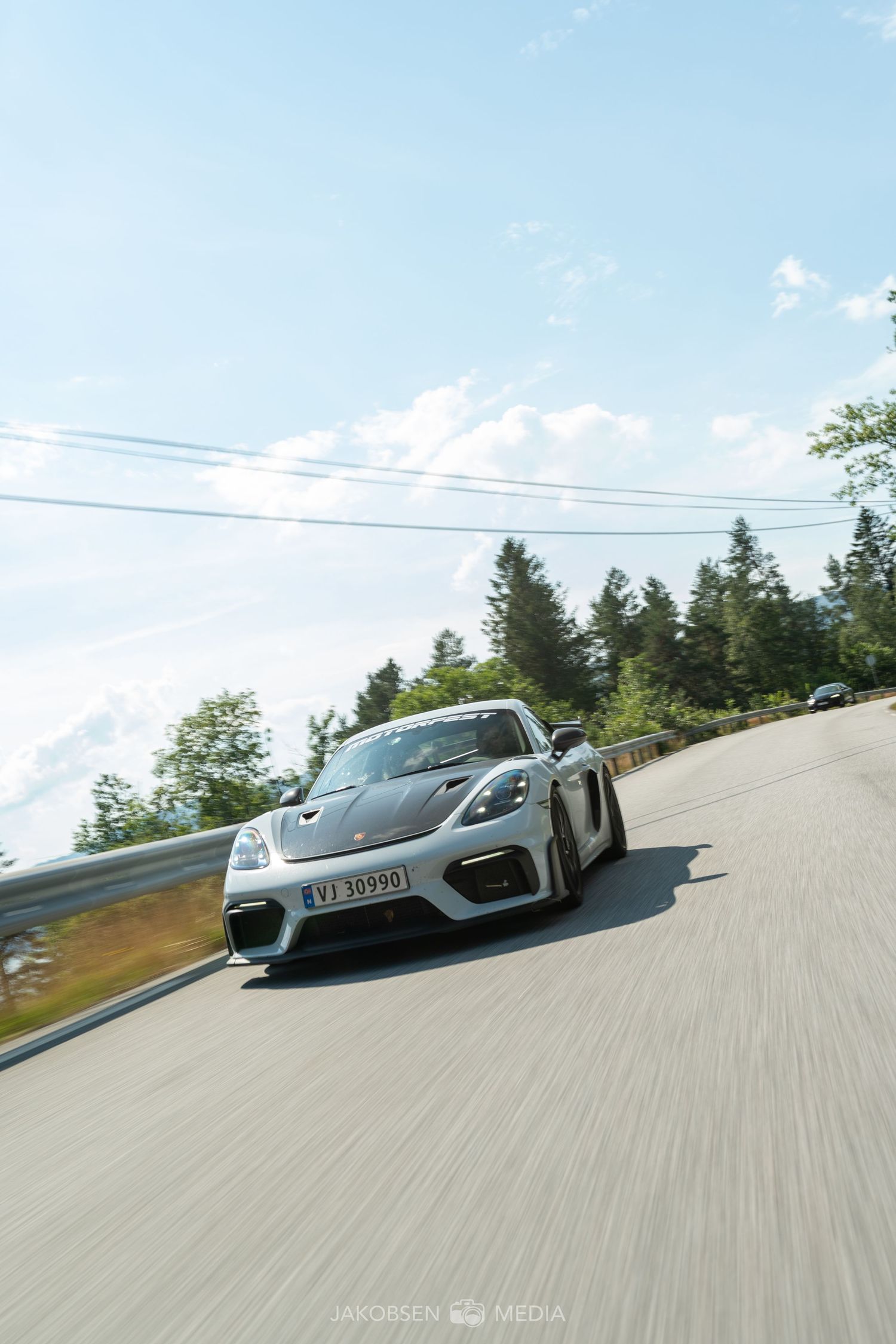 Silver sports car photographed in motion on a racetrack against blue sky and trees.