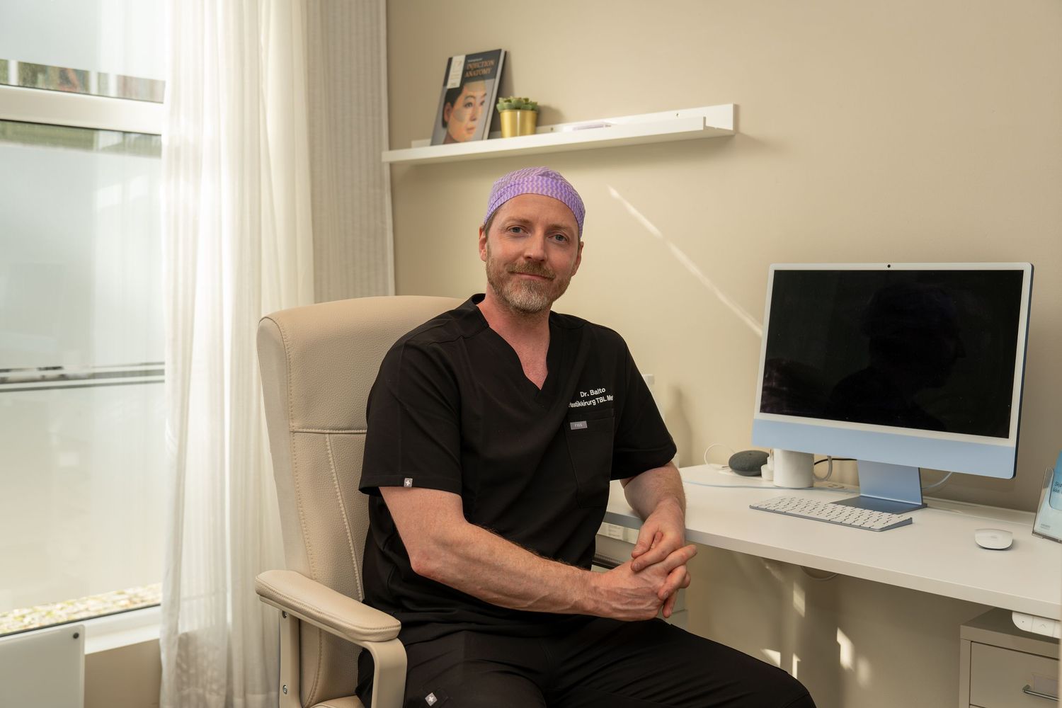 Man in black scrubs and a purple surgical cap sits in a beige chair beside a desk with an iMac in a bright, modern medical office.