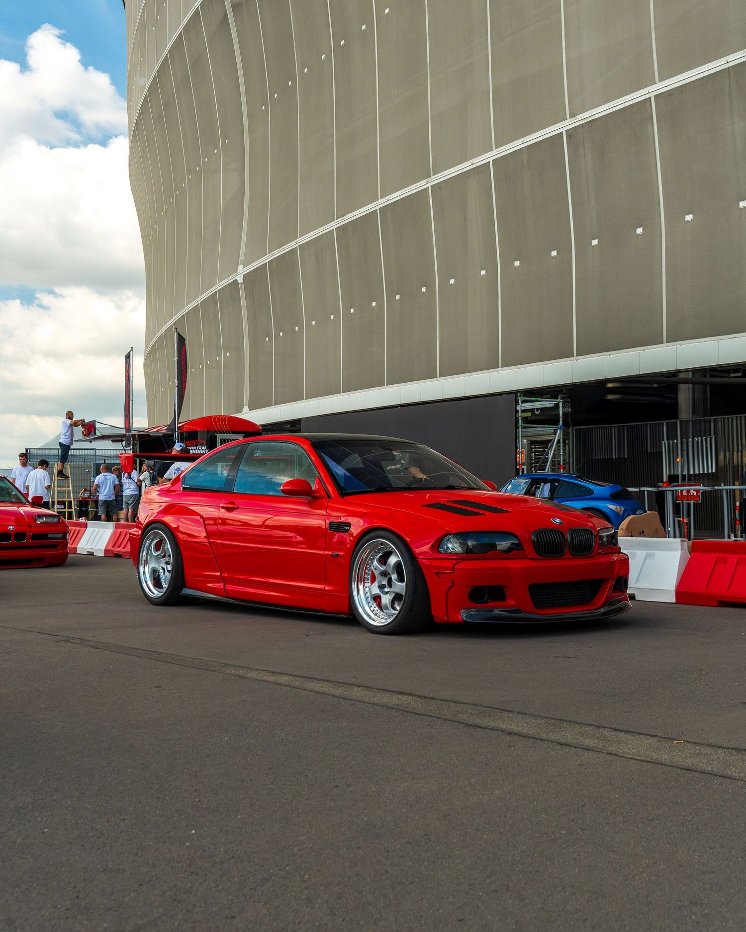 A bright red BMW M3 with lowered suspension and silver wheels parked outside a curved stadium structure.