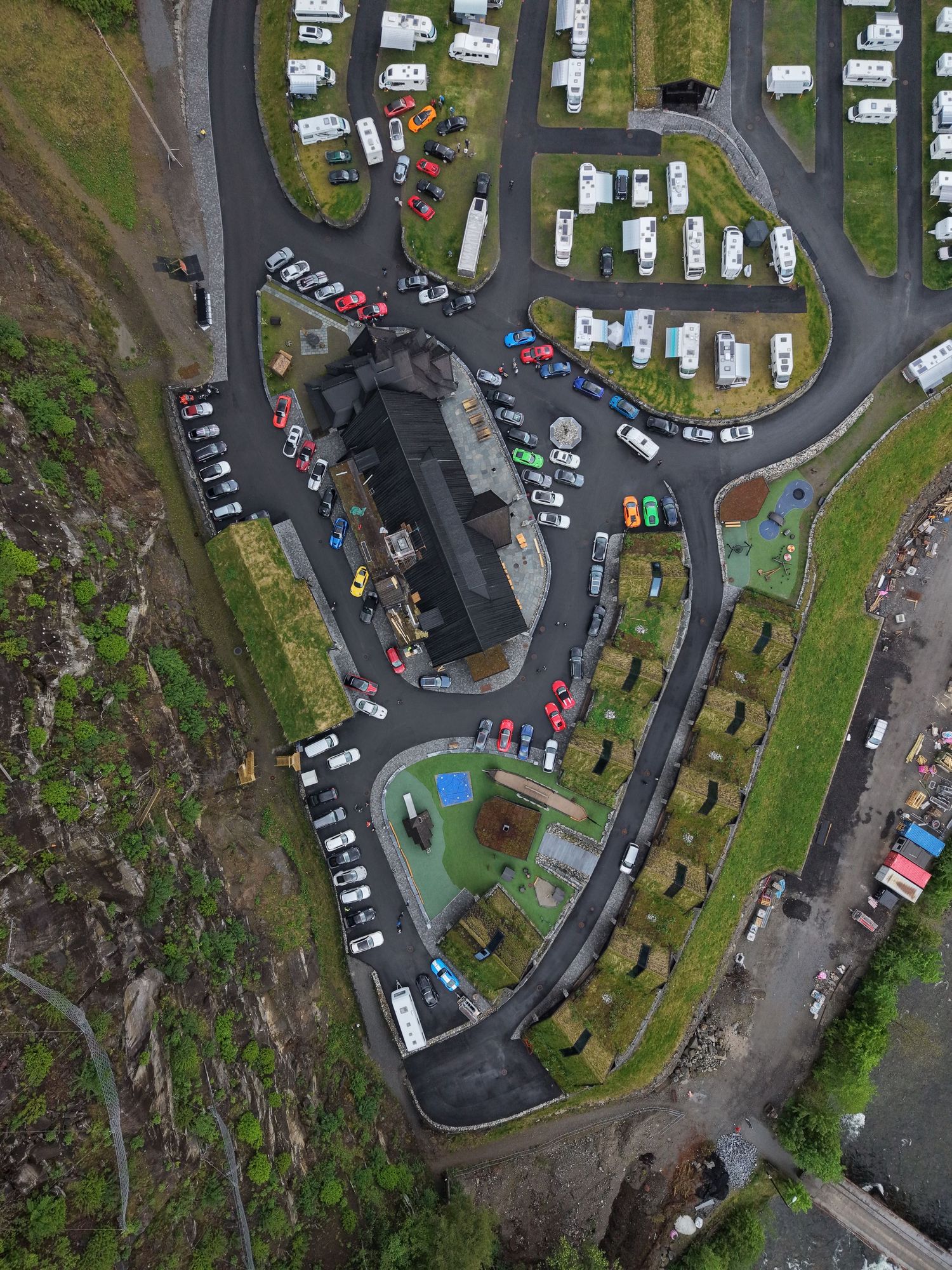 Aerial view of a hillside campground with parked cars, motorhomes and caravans along a winding road beside a dark-roofed building.
