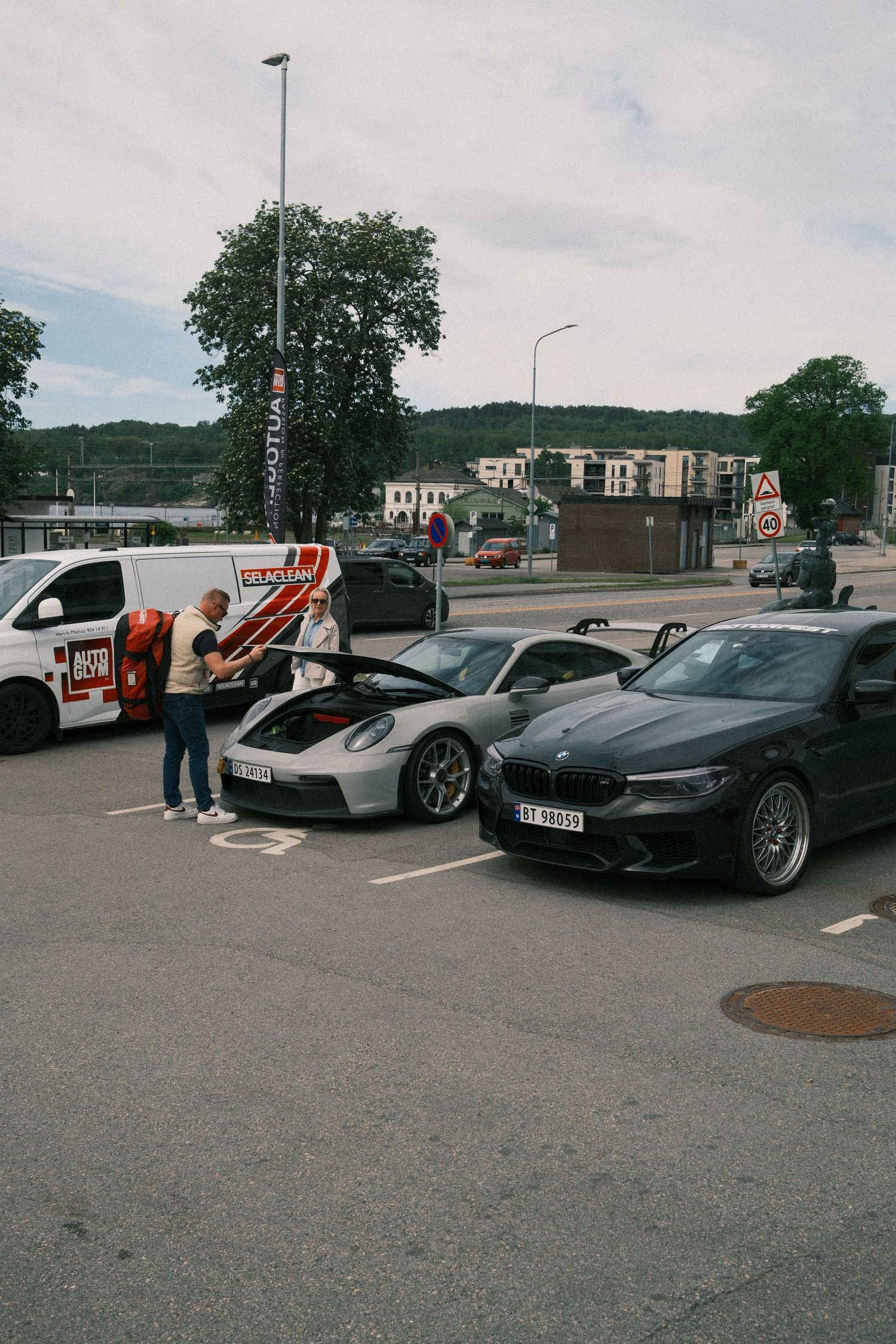 Two men inspect a silver sports car with its front hood open beside a black BMW in a parking lot, with a white van nearby.