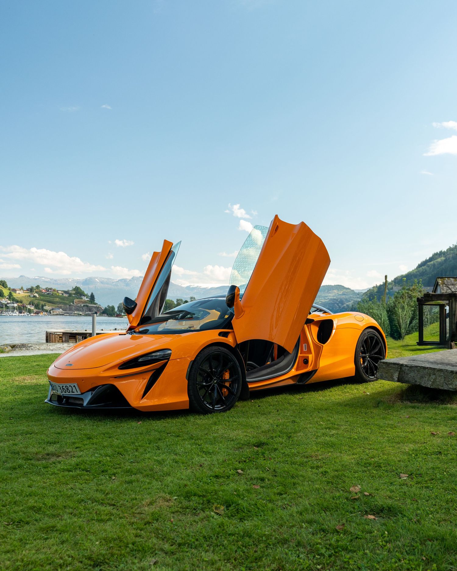Orange McLaren supercar with butterfly doors open parked on a green lawn beside a lake with mountains in the background.