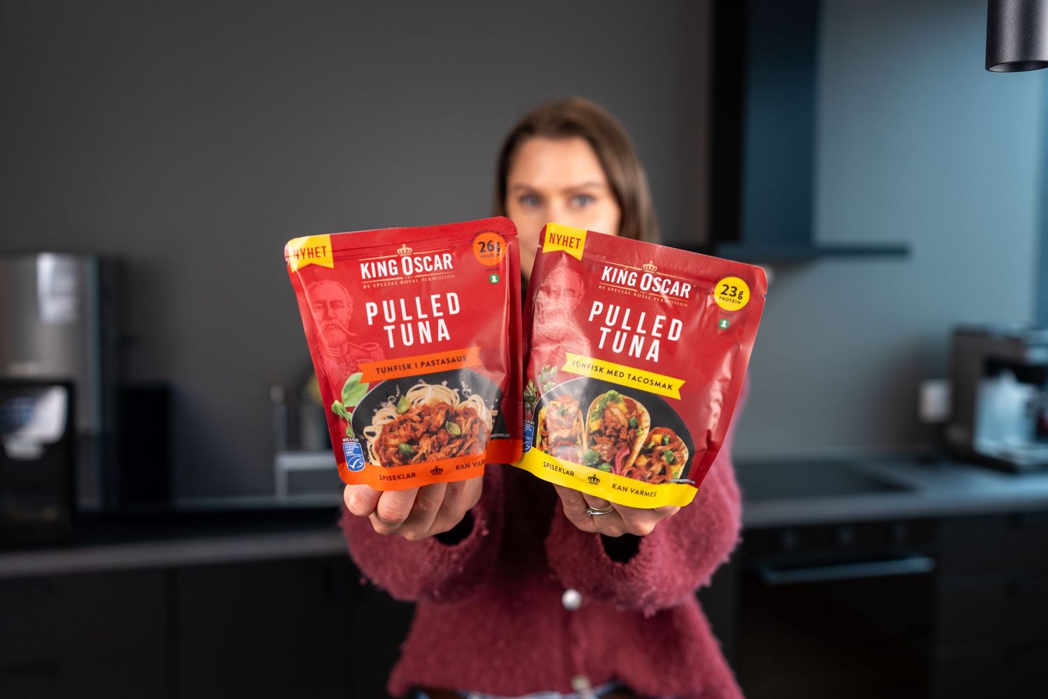 Woman holding two King Oscar Pulled Tuna pouches in a kitchen, showcasing different flavor varieties.
