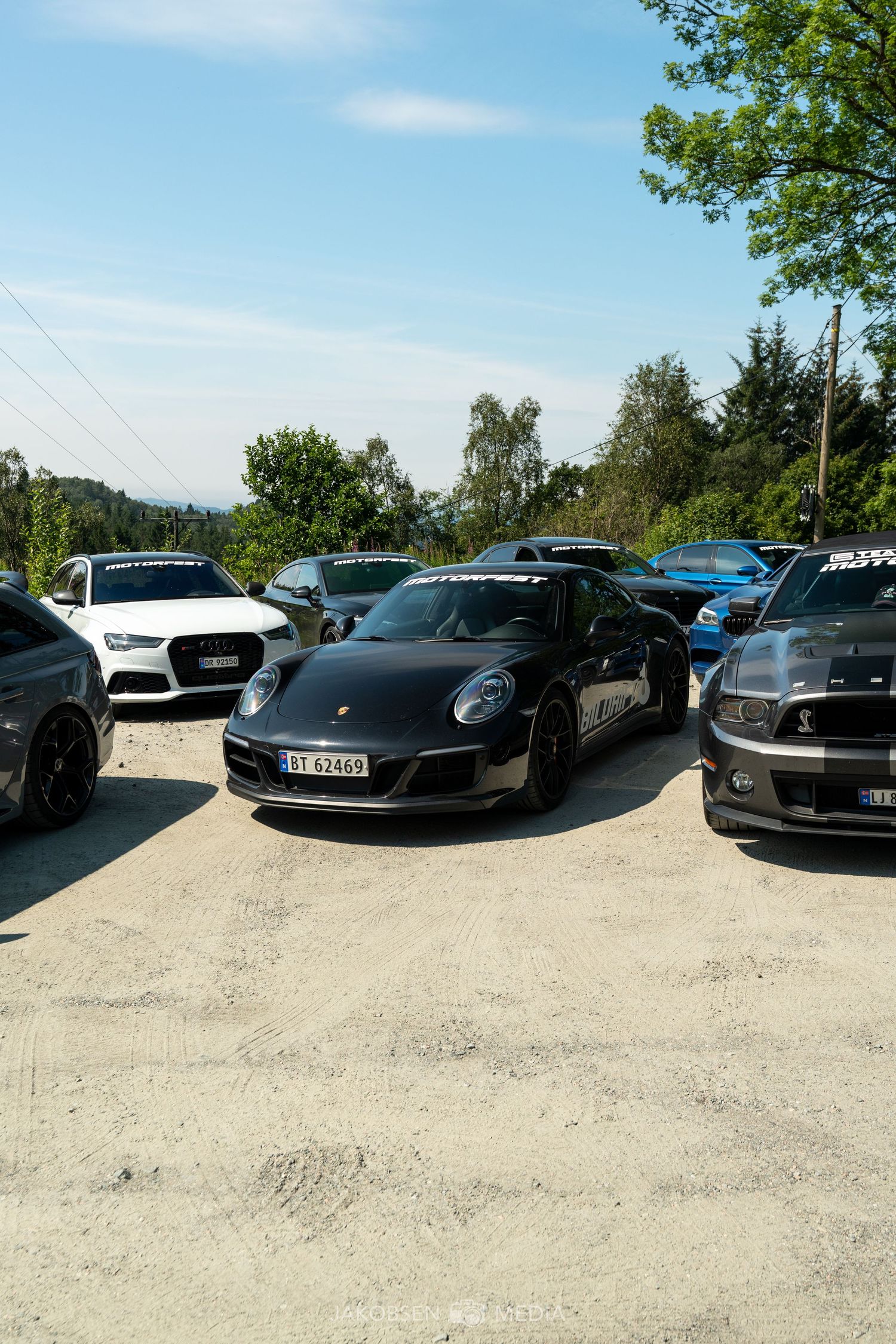 Matte black Porsche 911 parked among other luxury vehicles in a gravel parking lot.
