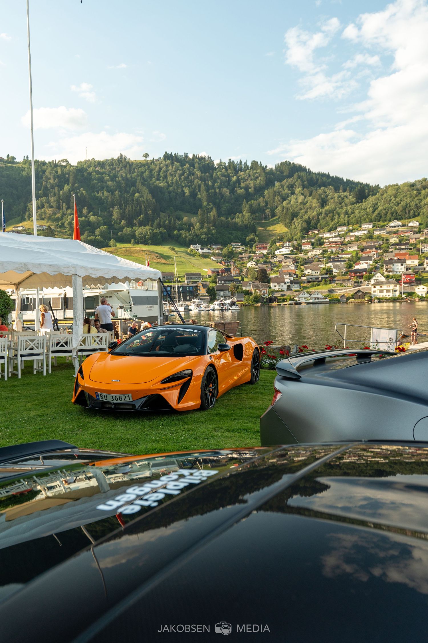 Orange McLaren sports car parked by a scenic lake vista with mountains and houses in the background.