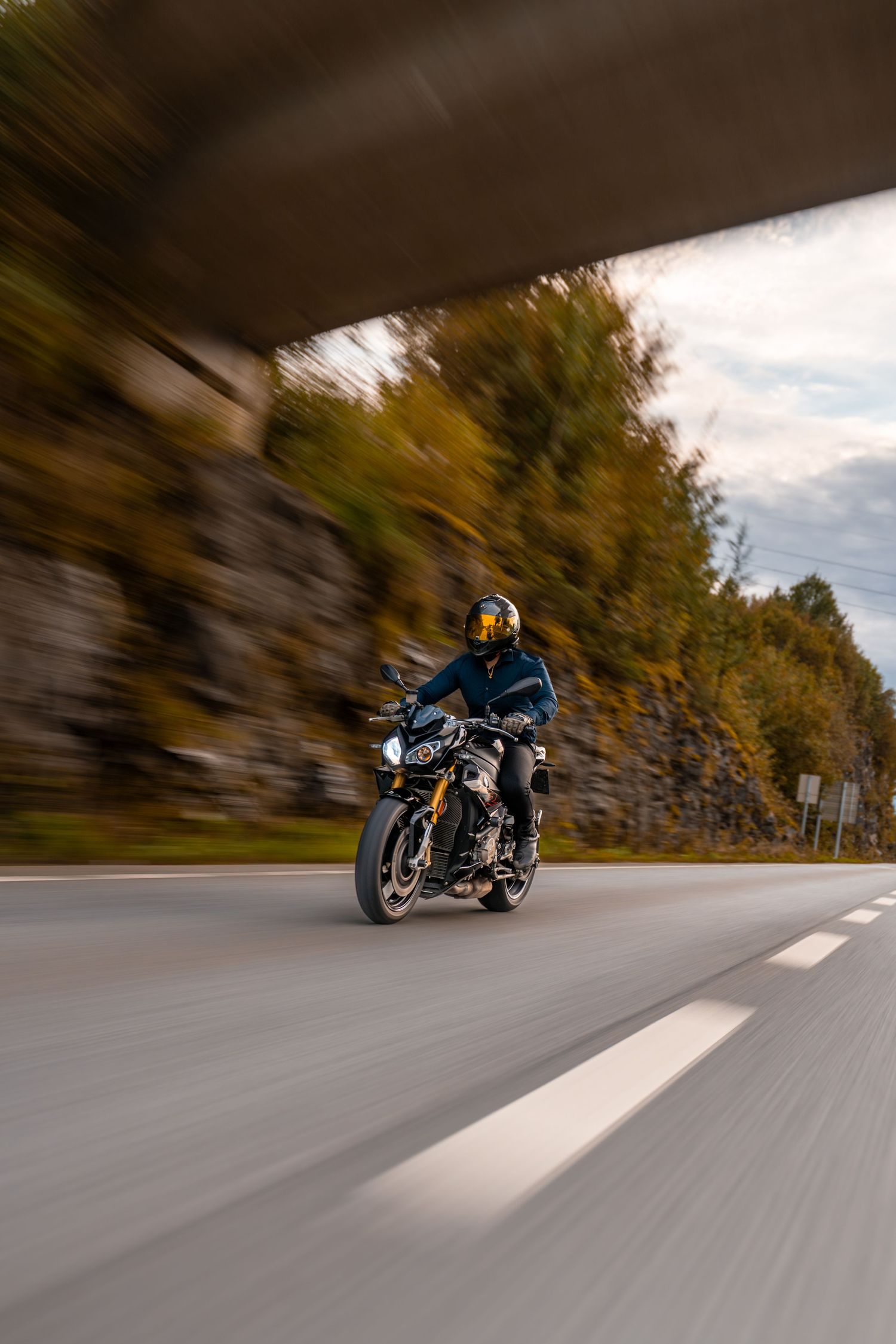 A motorcycle rider speeds down a winding road creating a dynamic motion blur effect in the autumn scenery.