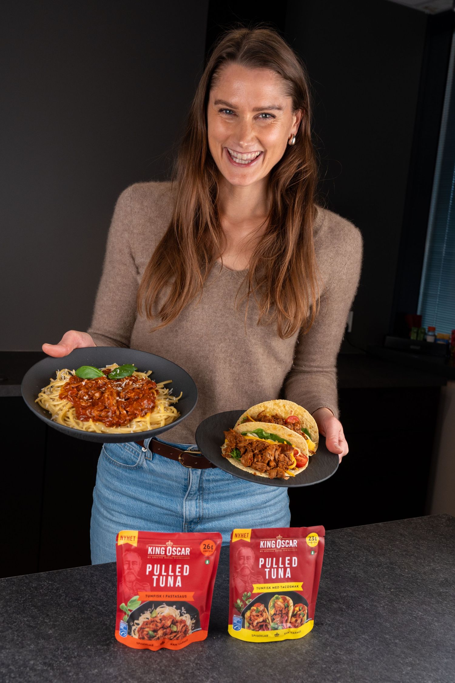 Smiling woman holds plates of spaghetti with pulled tuna sauce and pulled tuna tacos beside two King Oscar Pulled Tuna pouches.