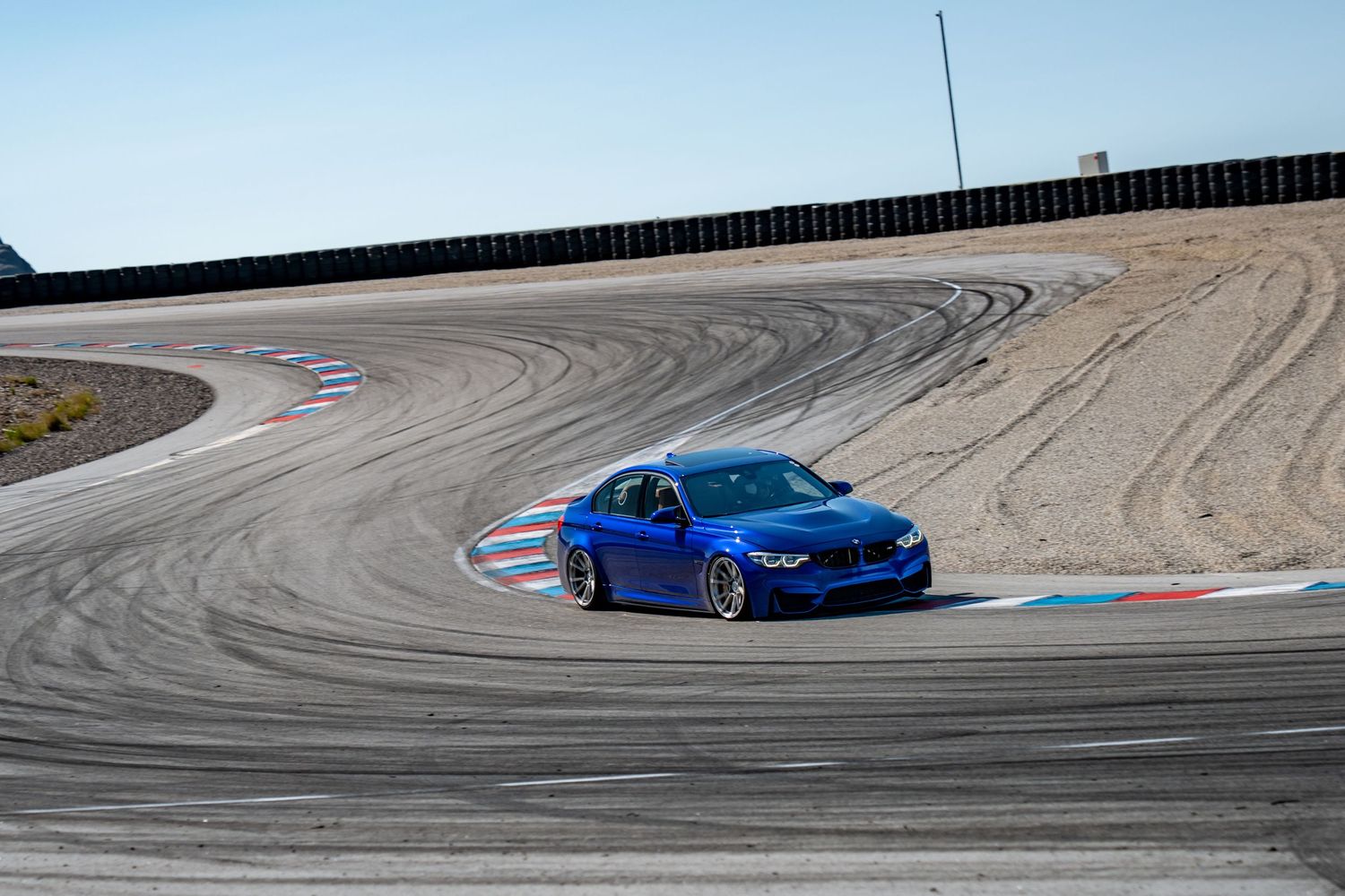 Blue BMW M3 sports car navigates a winding racetrack curve against a desert mountain landscape.