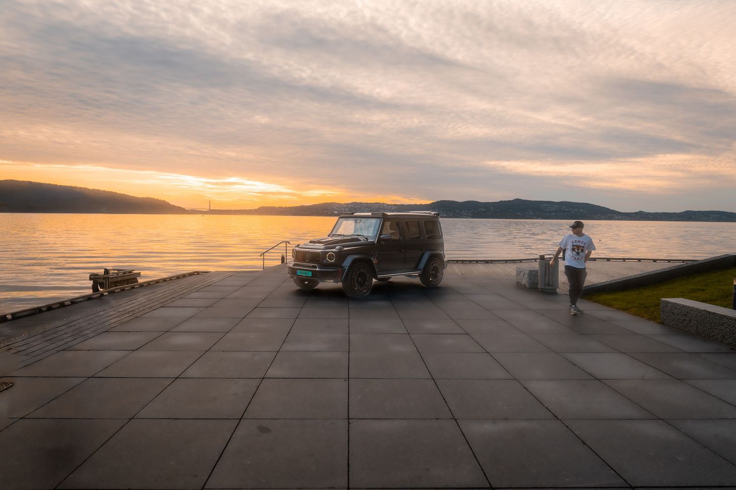 Panoramic view of a sunset over a waterfront parking lot with silhouettes of cars and mountains in distance.