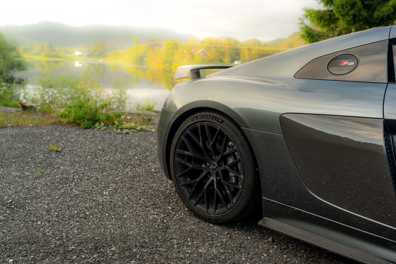 Silver sports car with black alloy wheels parked on gravel overlooking a lake during sunset.