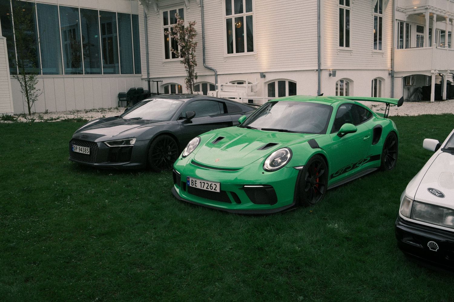 Green Porsche 911 GT3 RS with a rear wing parked on a lawn beside a dark gray Audi R8 in front of a white historic building.