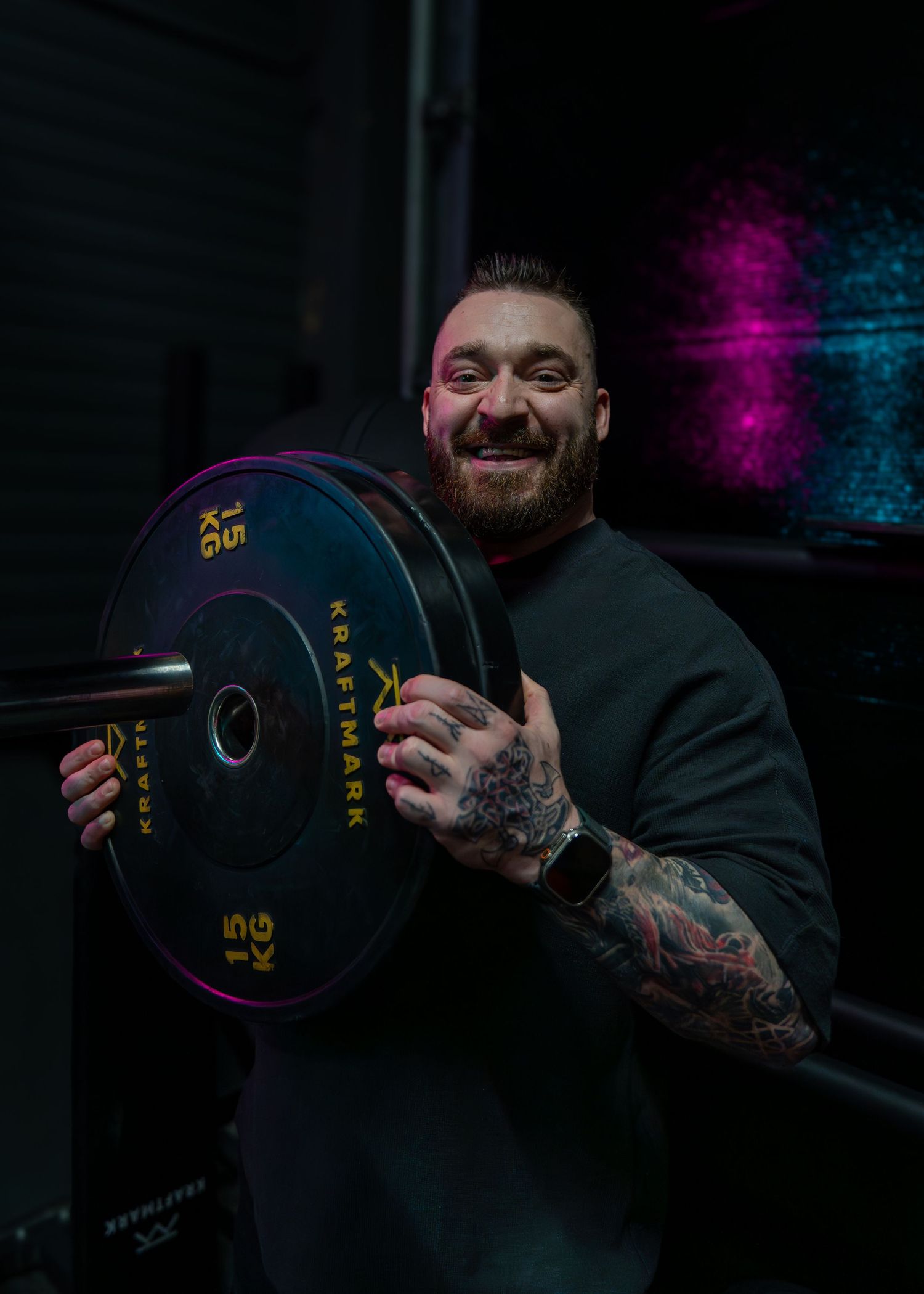 Strength athlete in black sweater holds a colorful Olympic weight plate in dramatic lighting with purple backdrop.