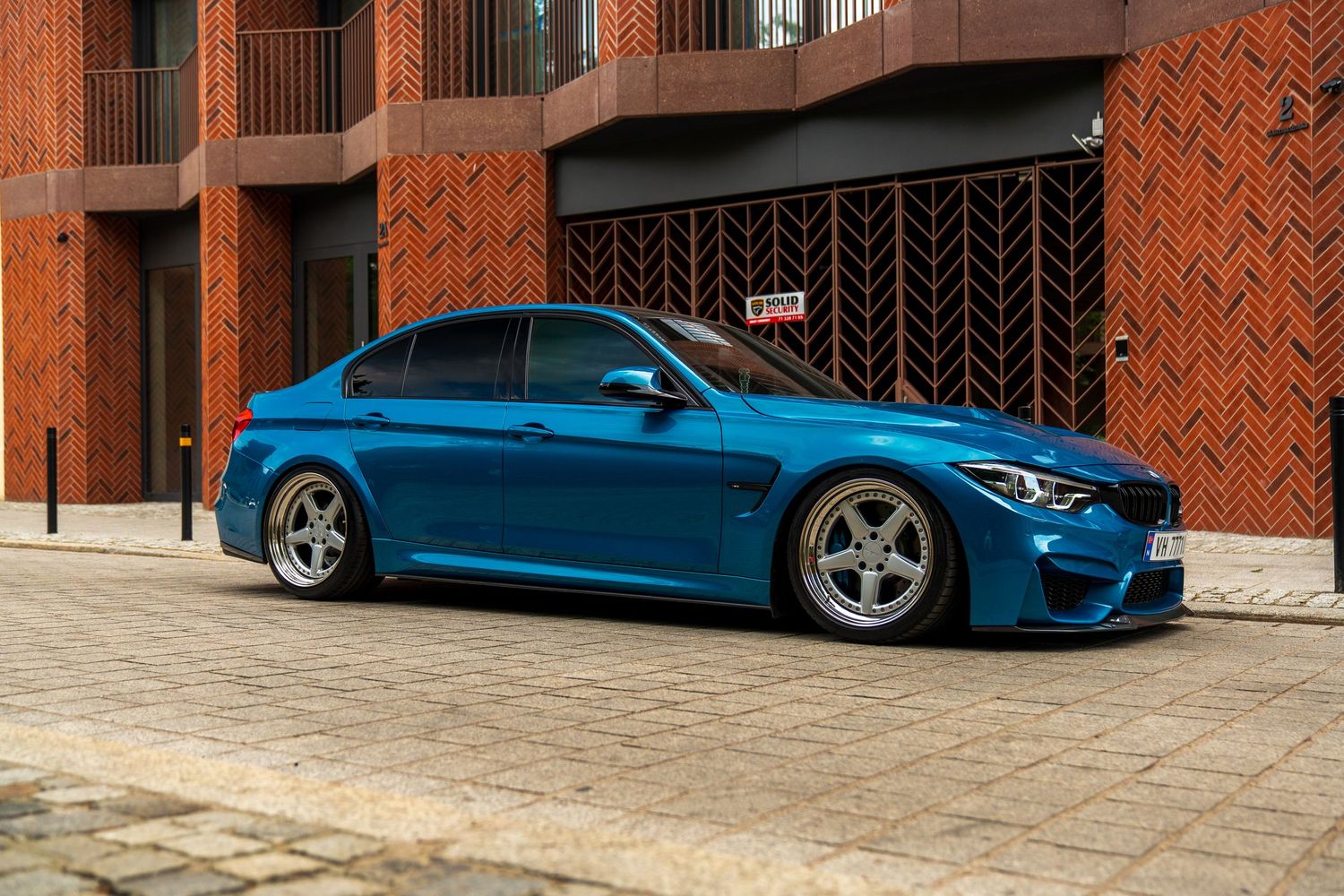 Modified BMW M3 in bright blue parked against a brick building with custom wheels and lowered suspension.
