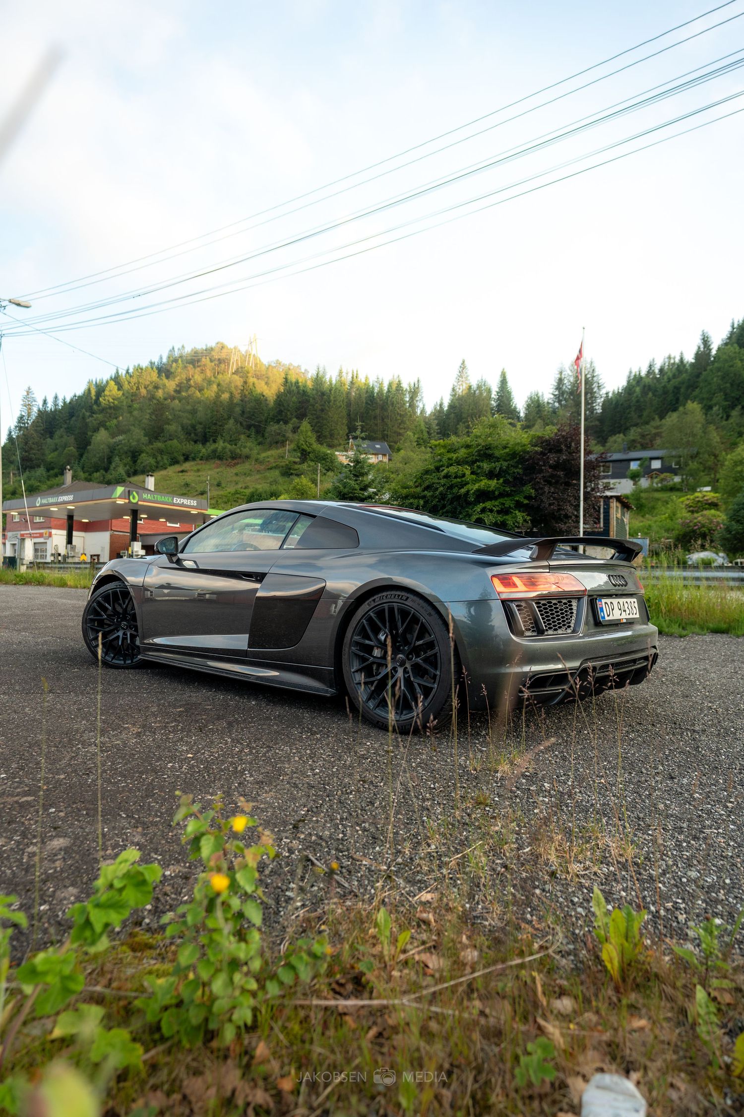 Gray luxury sports car with sleek design parked on gravel lot with power lines and forest in background.