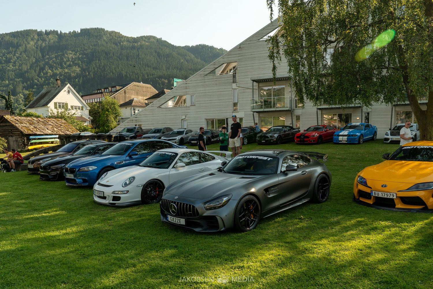 Panoramic view of luxury sports cars parked on grass with mountains and buildings in the background.