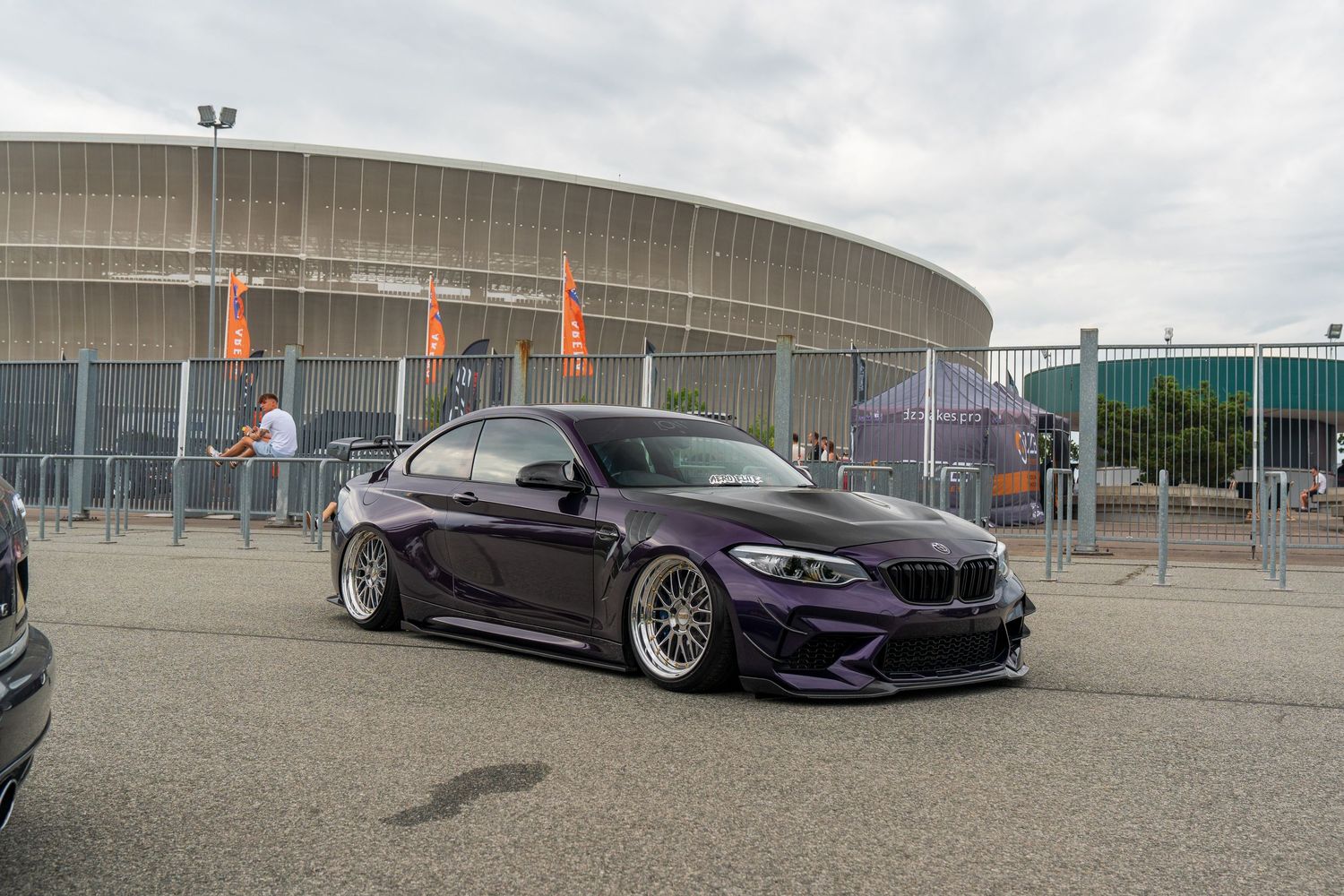 Purple BMW M2 with lowered suspension and custom wheels parked in front of a large arena building.
