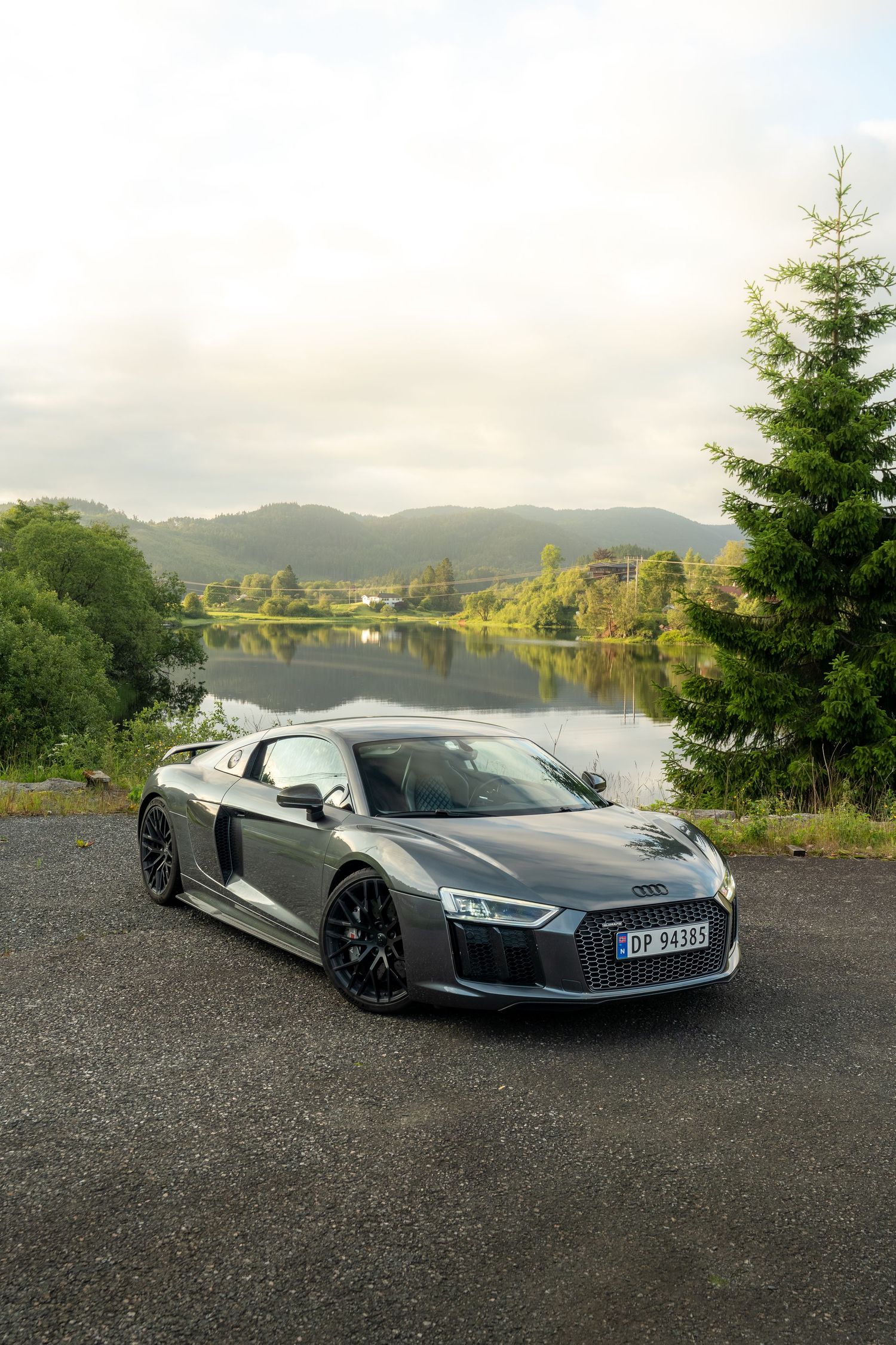 Grey Audi R8 supercar photographed in front of a scenic lake and mountain landscape.