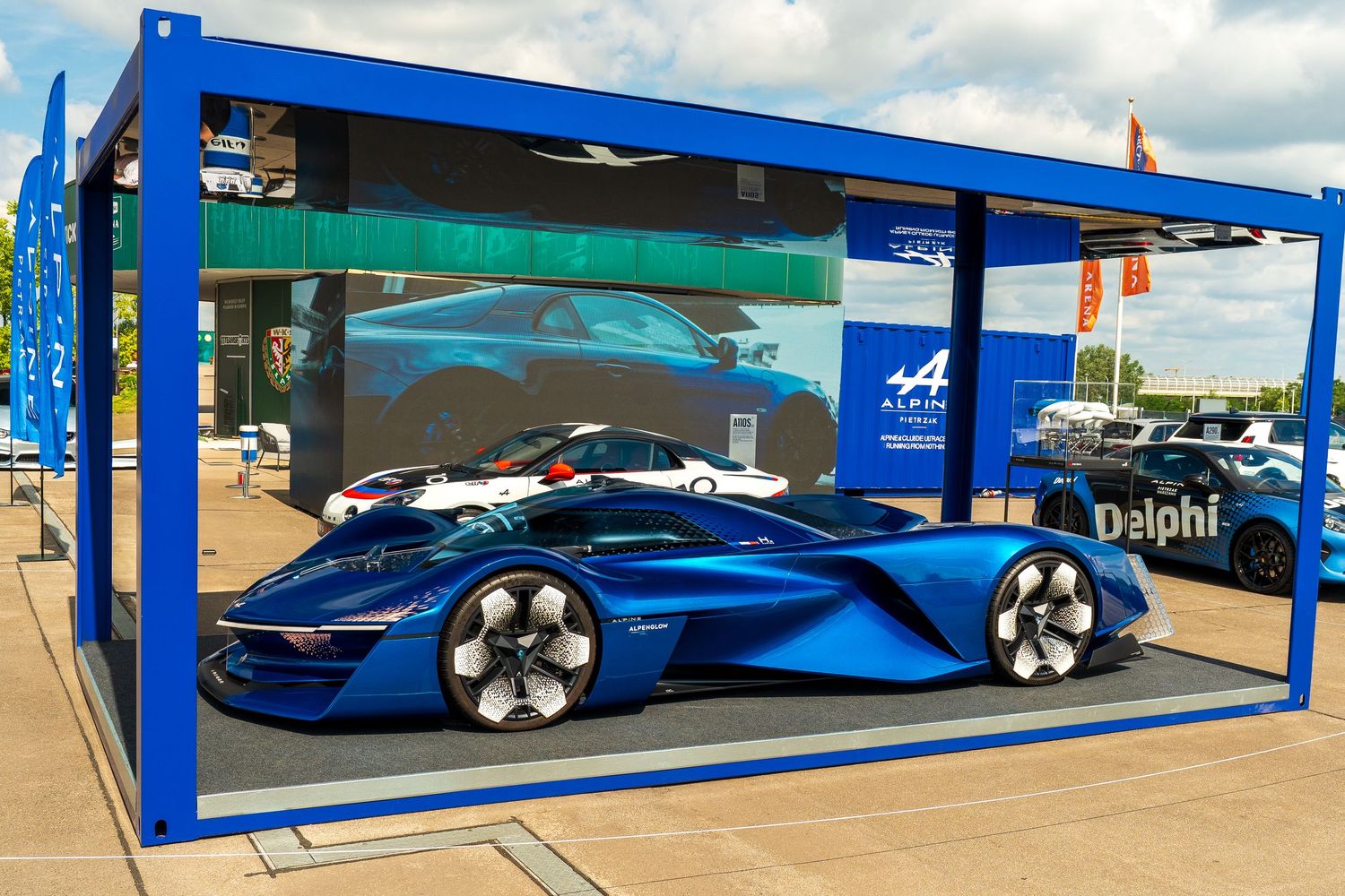 A sleek blue concept car with futuristic styling displayed under a blue exhibition booth canopy at an outdoor event.