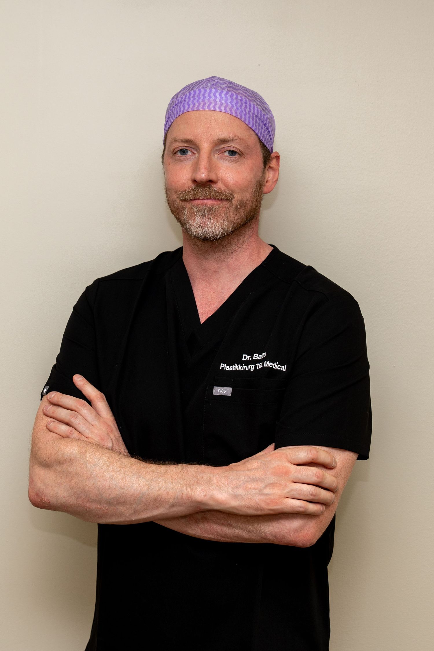 Smiling surgeon with arms crossed wearing black scrubs with embroidered name and a purple patterned surgical cap against a neutral wall.