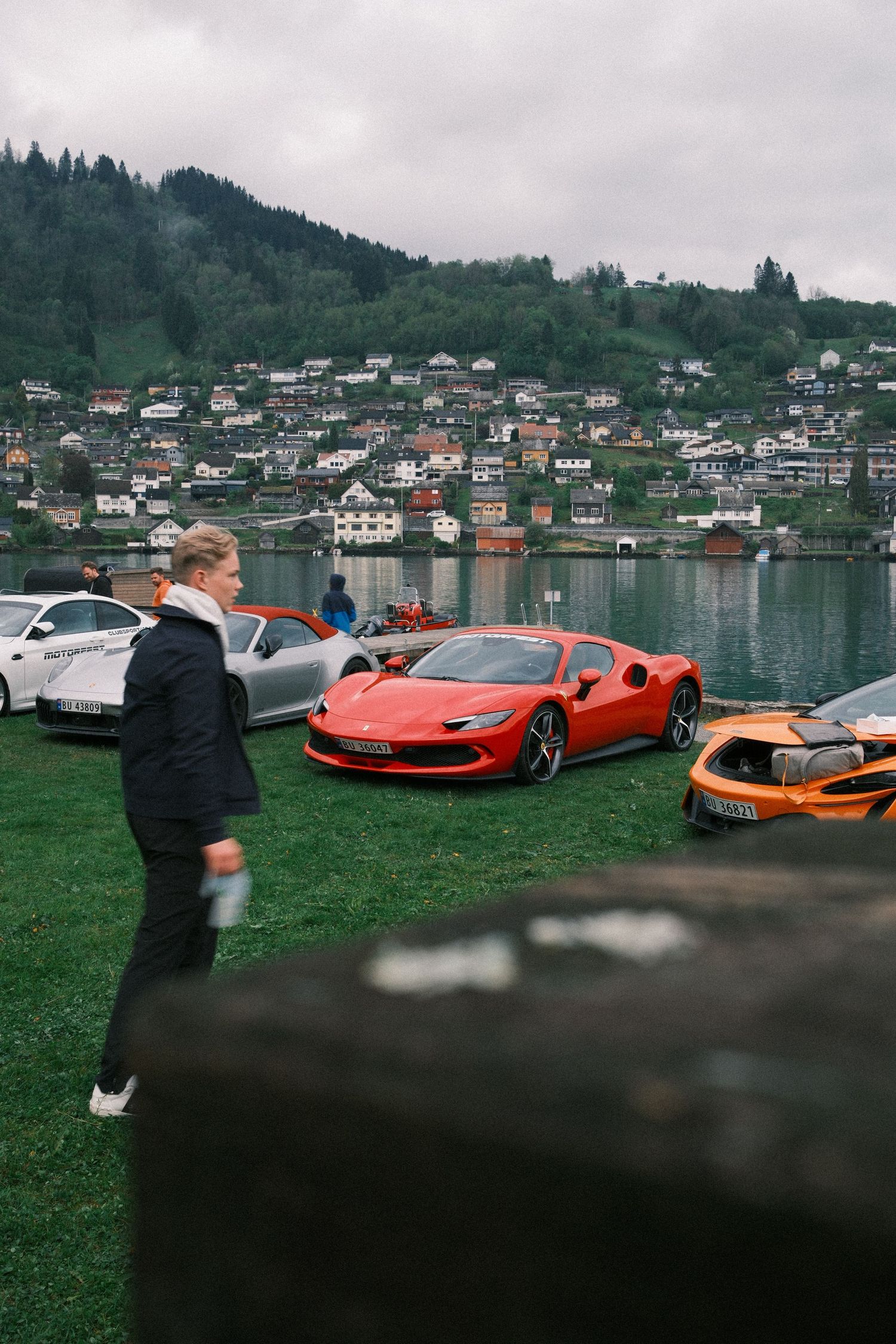 Red Ferrari sports car parked on a grassy lawn beside an orange supercar, with a lakeside village and forested hills in the background.