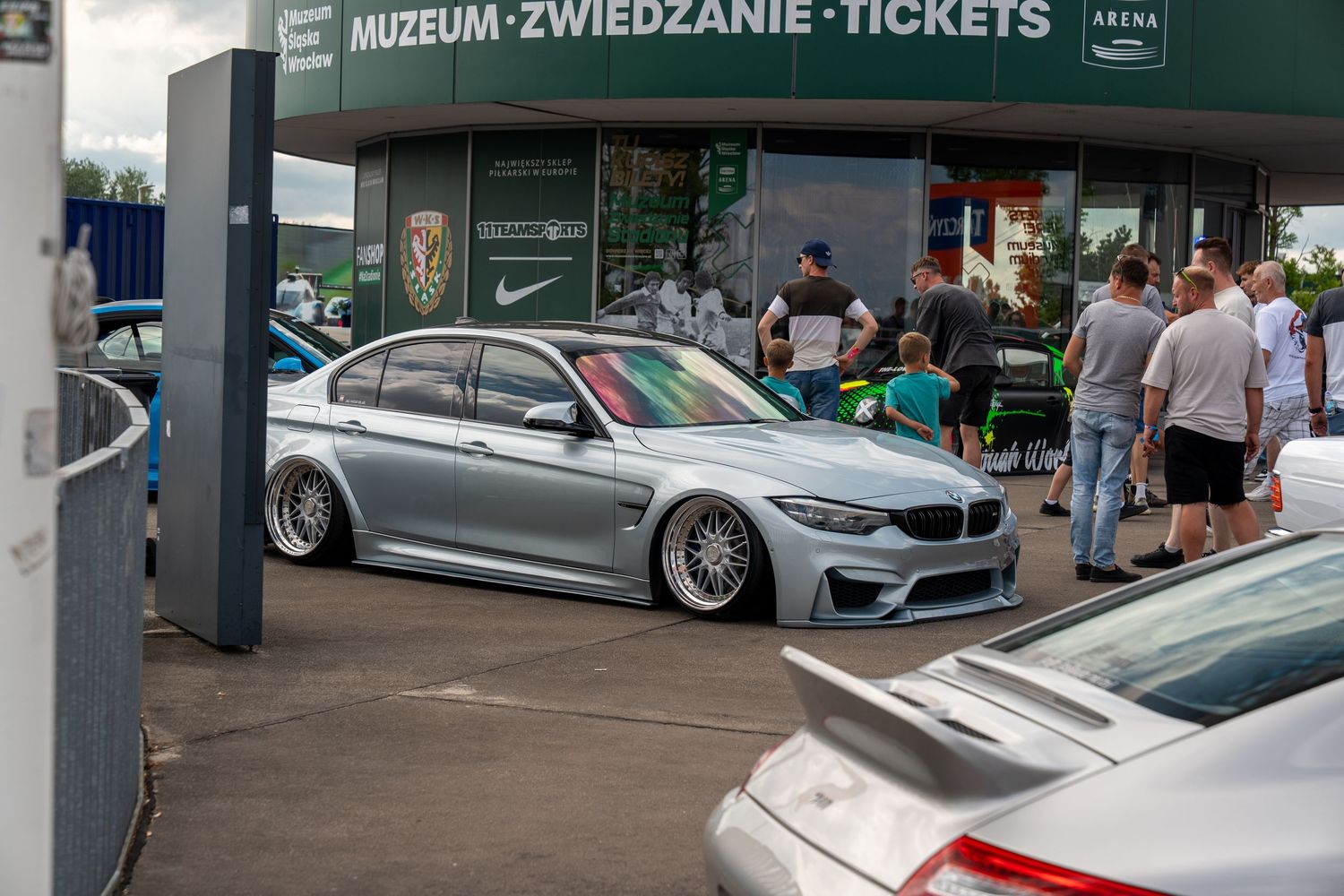 A lowered silver BMW M3 with aftermarket wheels parked outside a museum entrance.