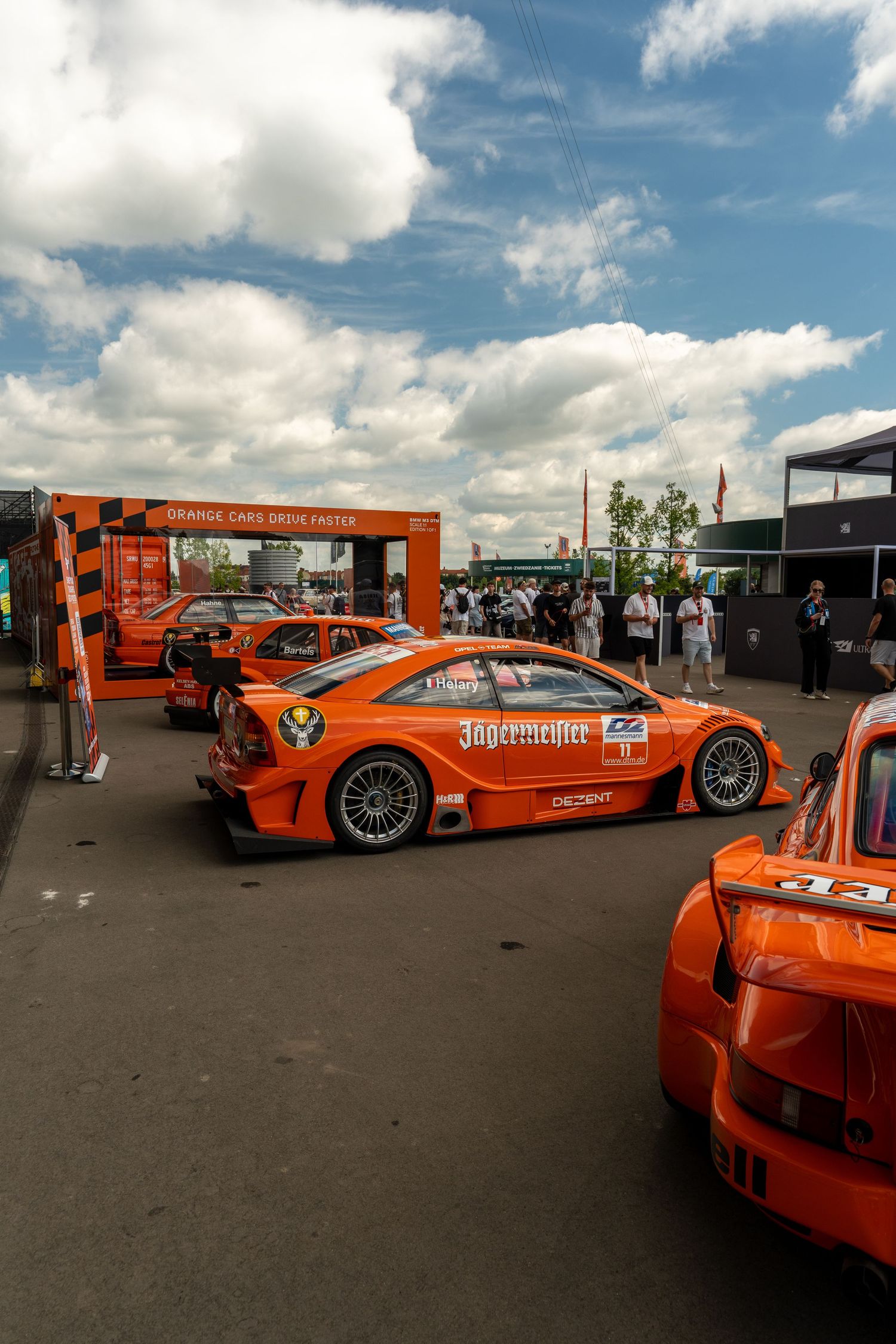 Orange race cars lined up at an outdoor motorsport event under cloudy blue skies.