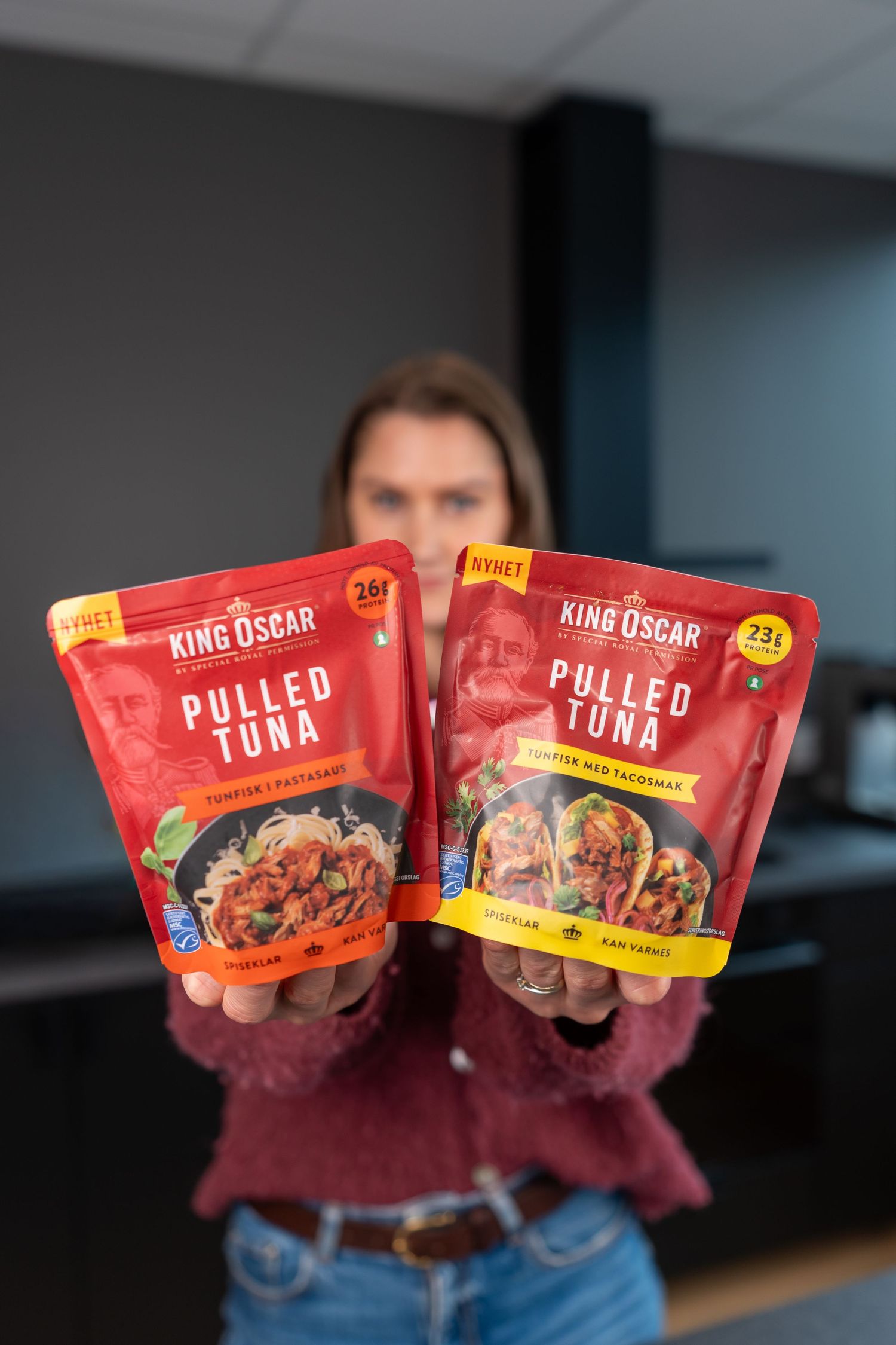 Person holding two red King Oscar Pulled Tuna pouches, one pasta-style and one taco-style, with 23g protein labels.
