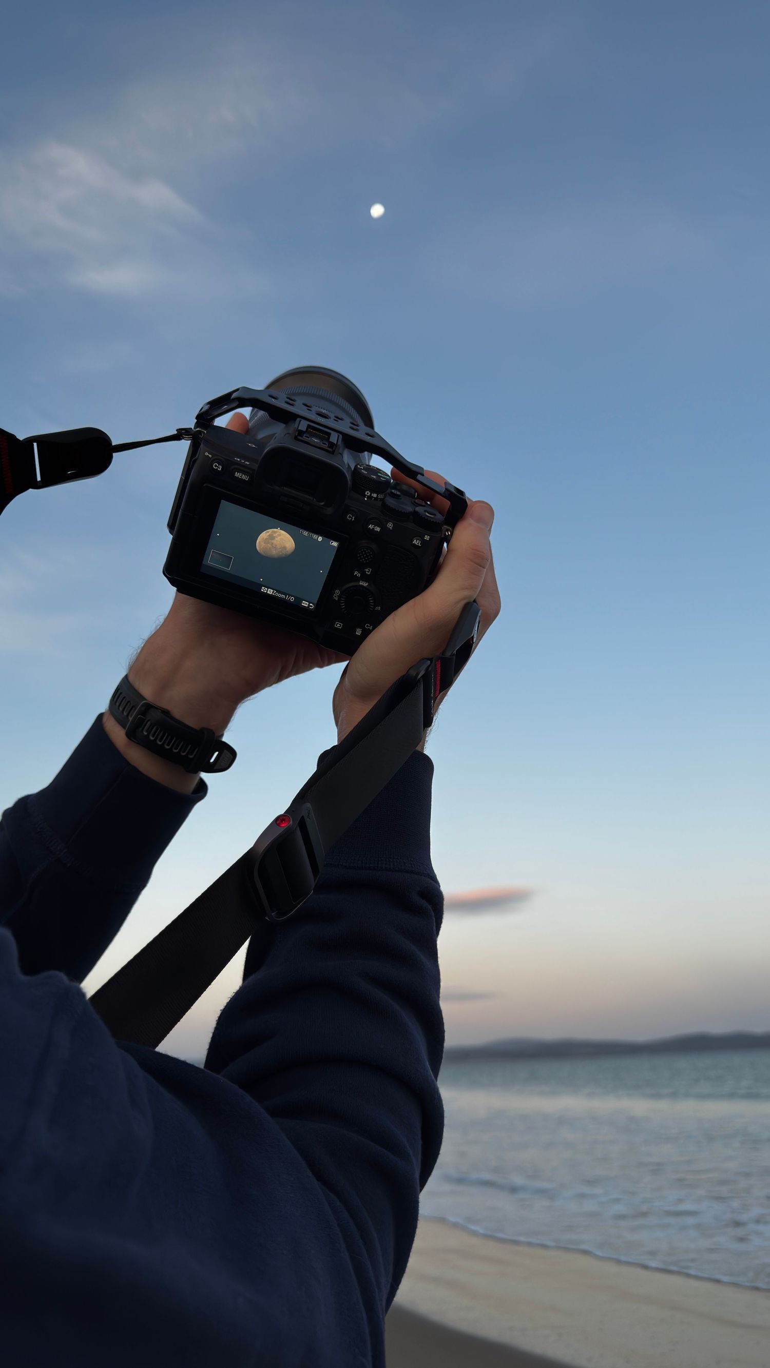Photographer holds camera up to capture the moon over a calm ocean at dusk on a beach.