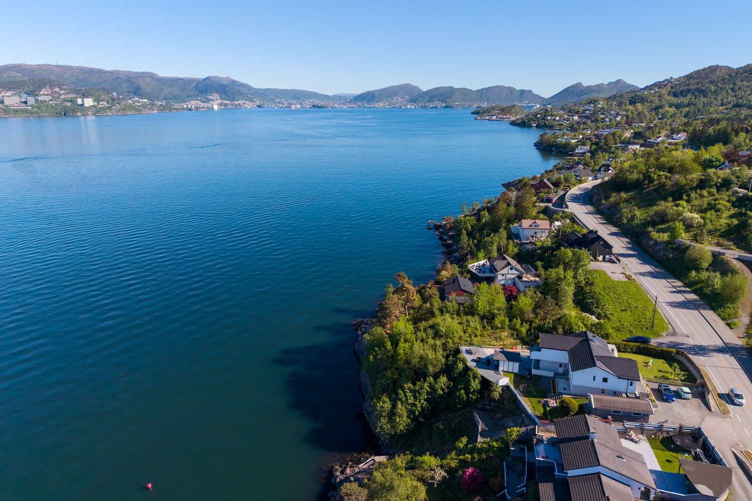 Aerial view of a deep blue fjord lined with waterfront homes, green hillsides, and distant mountains.