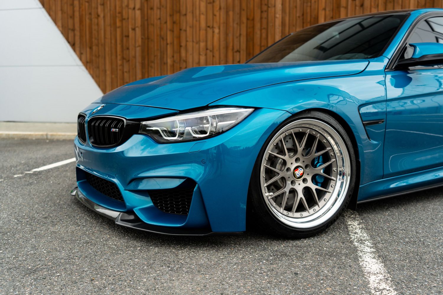 Close-up view of a bright blue BMW M4 featuring a carbon fiber front splitter and silver multi-spoke custom wheels.