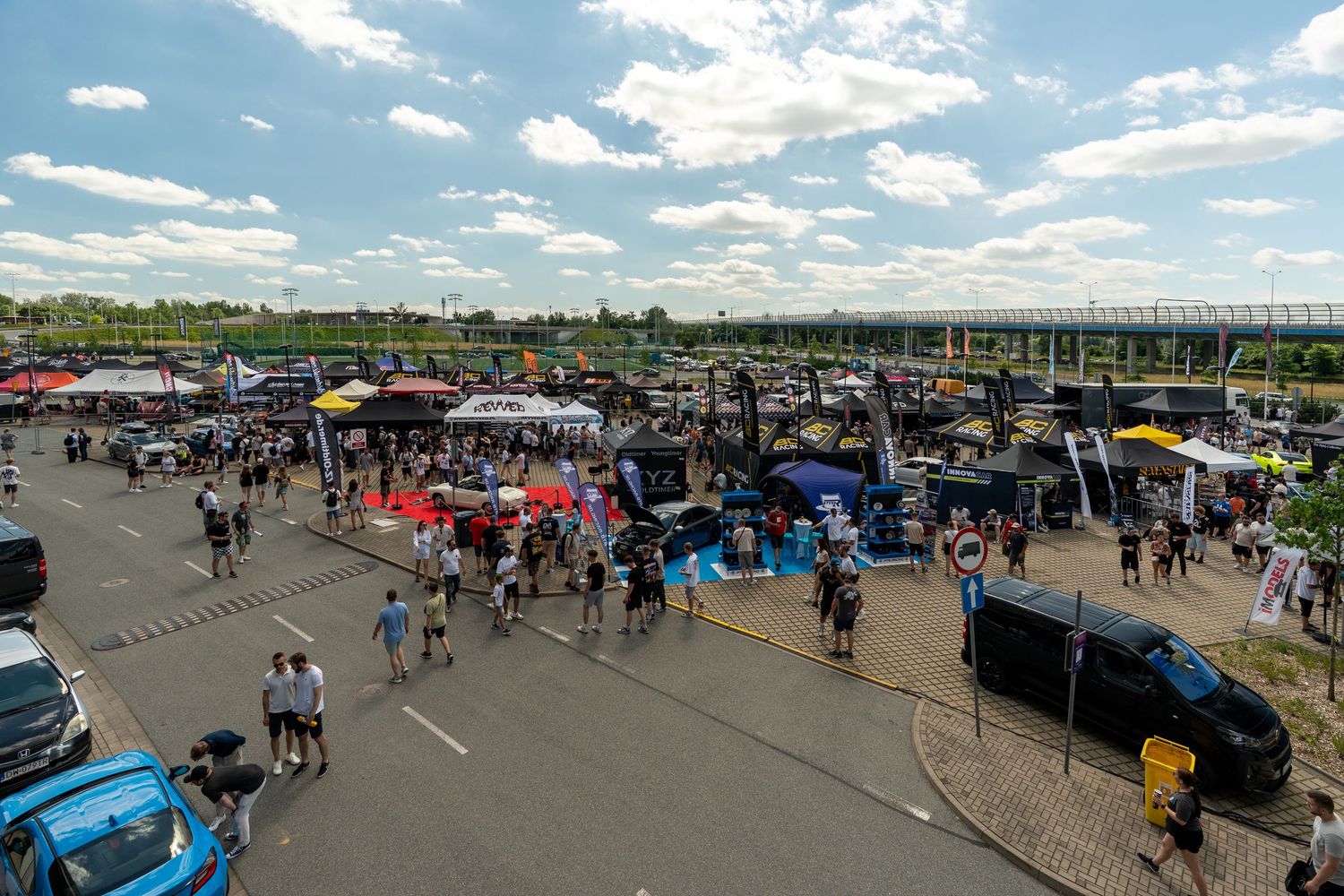 An aerial view of a crowded outdoor car show with vendor tents and spectators under blue skies.