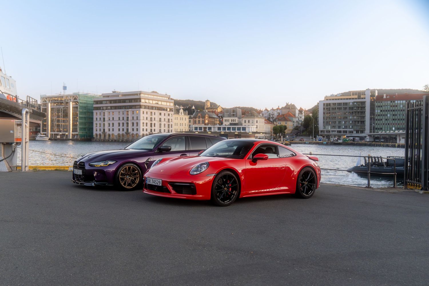 Three luxury sports cars including a bright red Porsche parked along a waterfront with city buildings in the background.