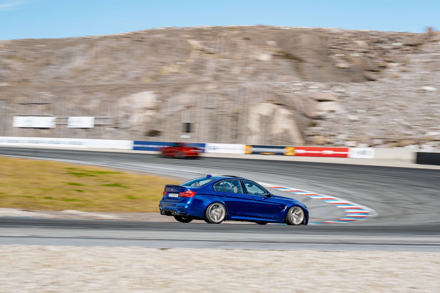 A blue sports car speeds through a racetrack turn during a motorsport competition on a sunny day.