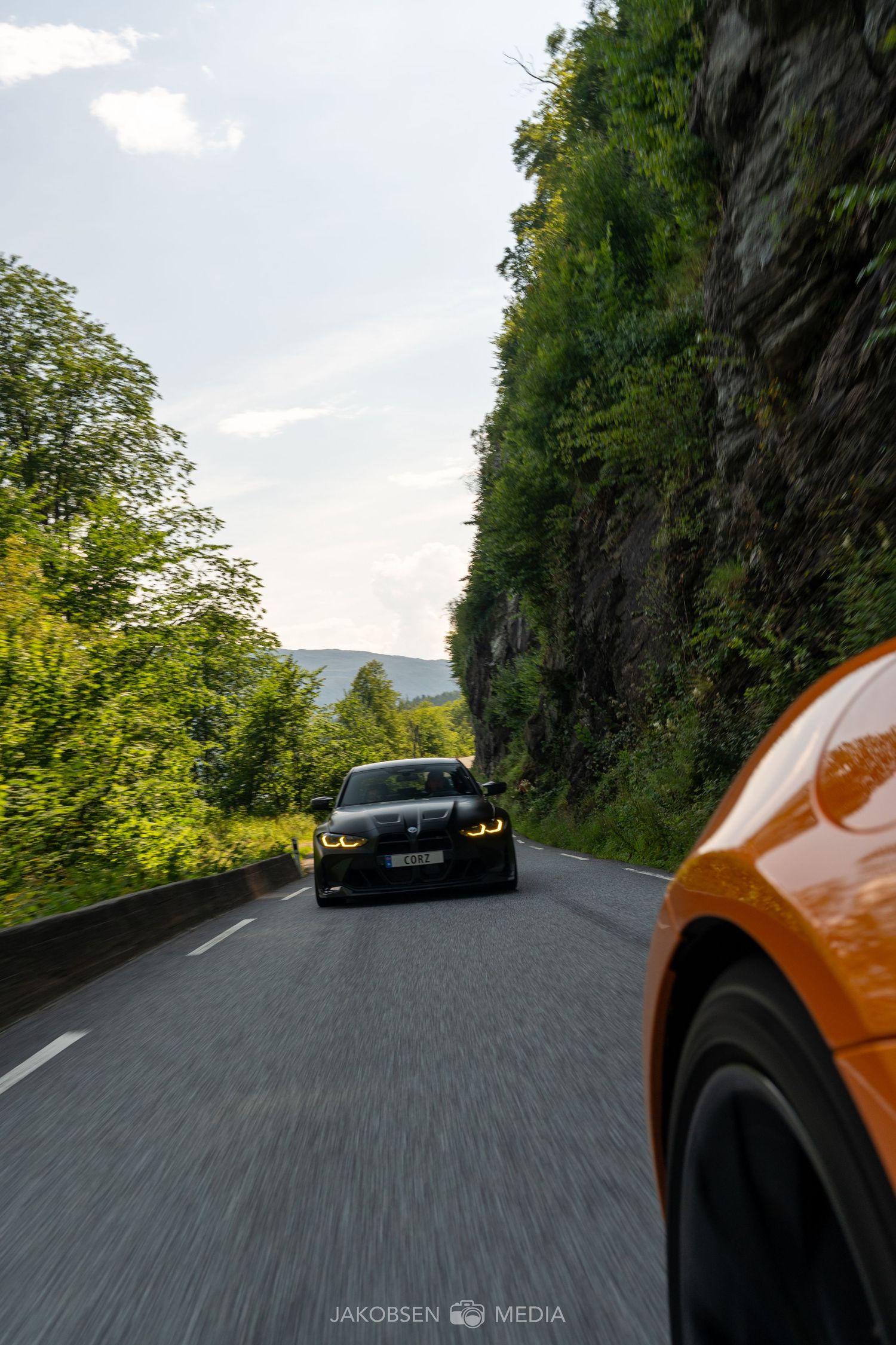 POV shot of driving on a winding mountain road with lush green cliffs and another vehicle ahead.