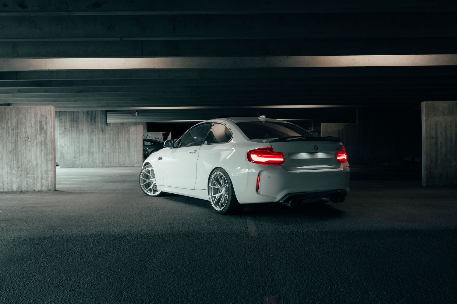 White BMW M2 sports car parked in dark underground garage with dramatic lighting.