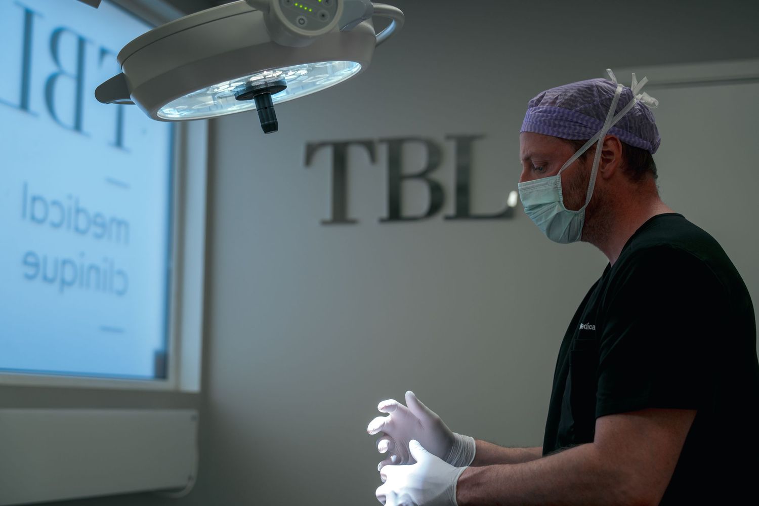 Surgeon in scrubs, purple cap and mask adjusts white gloves beneath an operating light at TBL medical clinique.