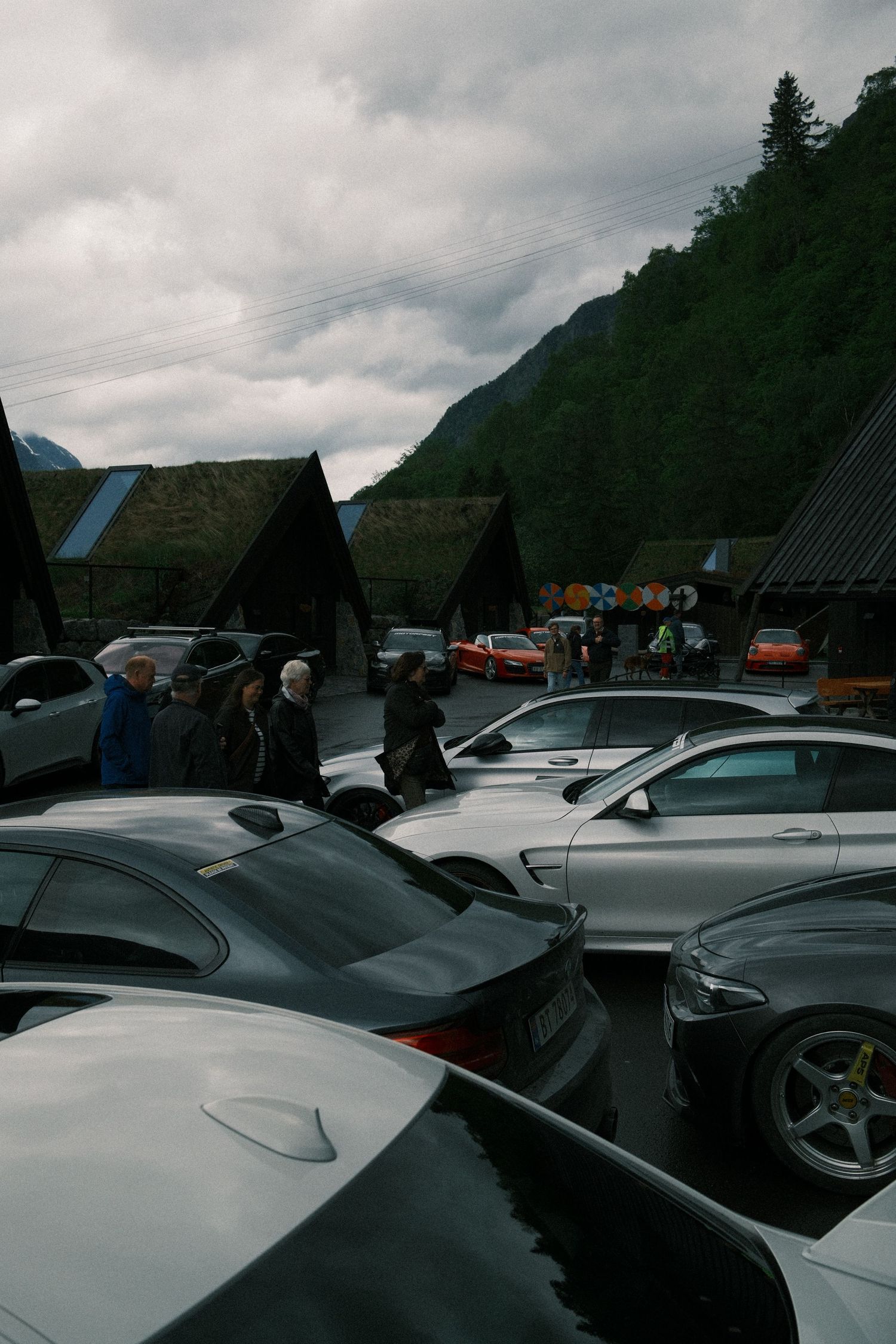 Cars parked in a wet lot as people gather near turf-roofed buildings, with a misty green mountain rising under a cloudy sky.