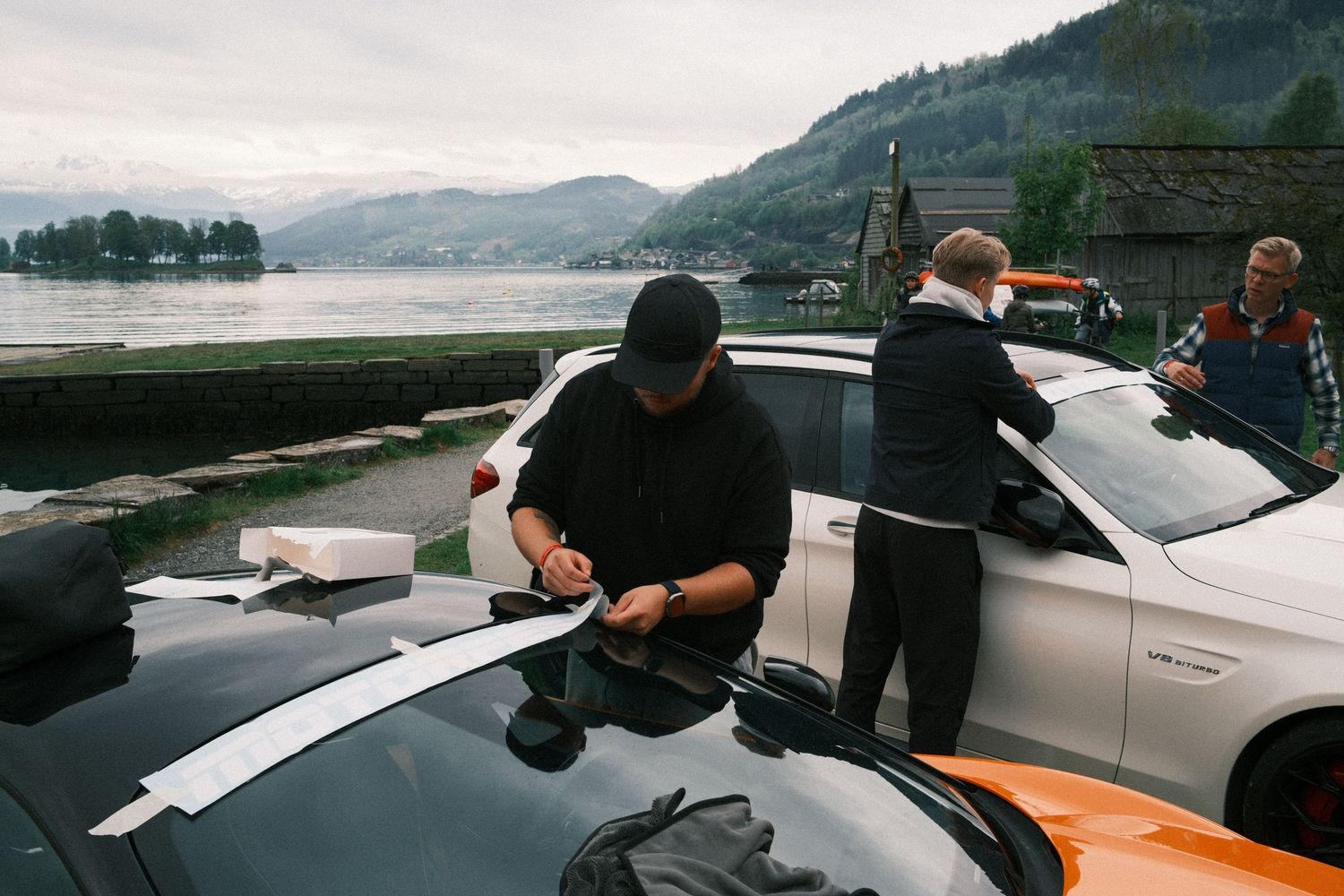 Three men gather around parked sports cars beside a calm mountain lake, one signing papers on a car hood on a cloudy day.